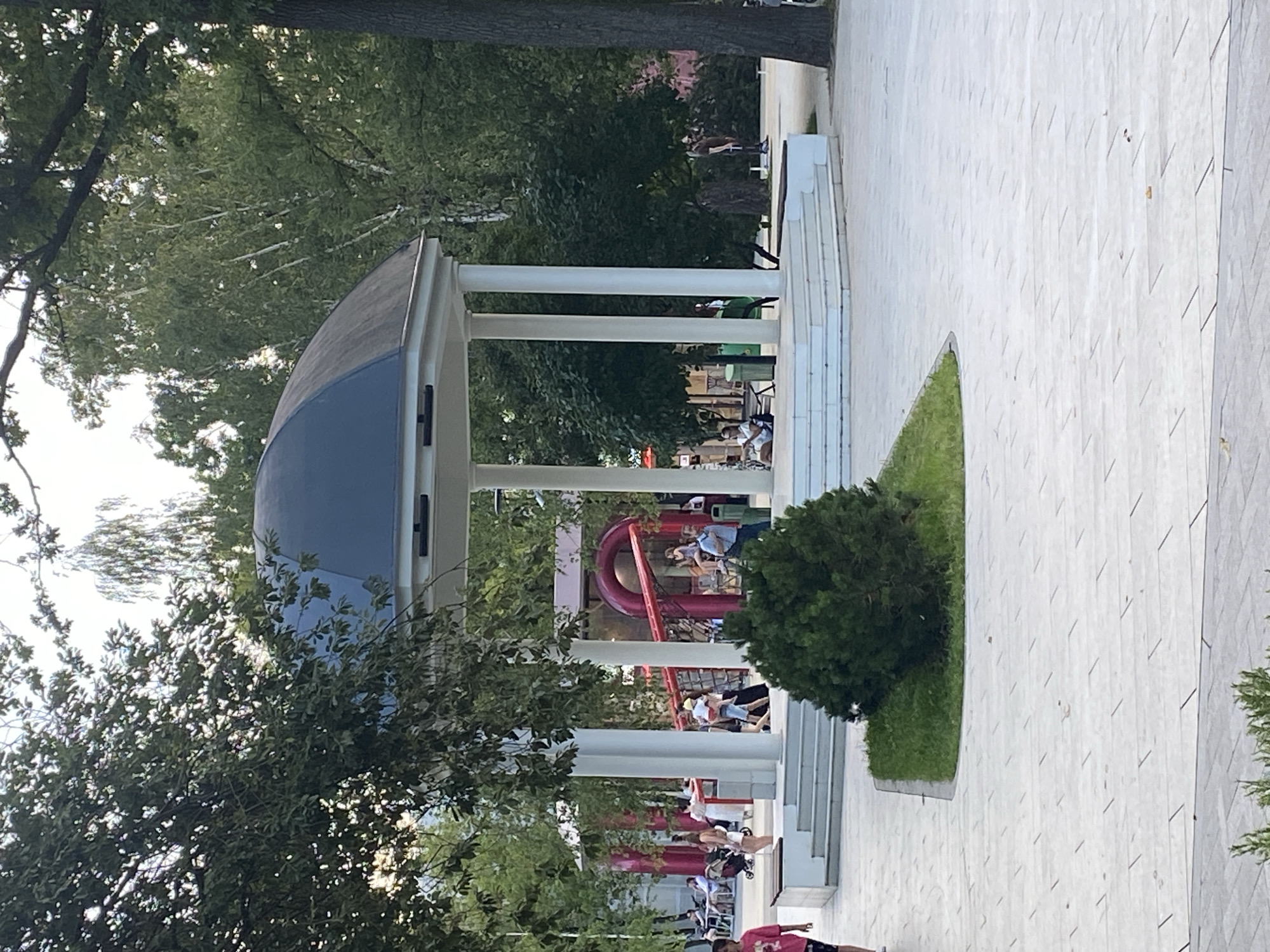 The image shows a public park with a gazebo and a playground in the background. People are visible walking, sitting, and interacting in the park. The environment appears to be urban, with trees and paved pathways. The overall scene is a snapshot of daily life in a city park.