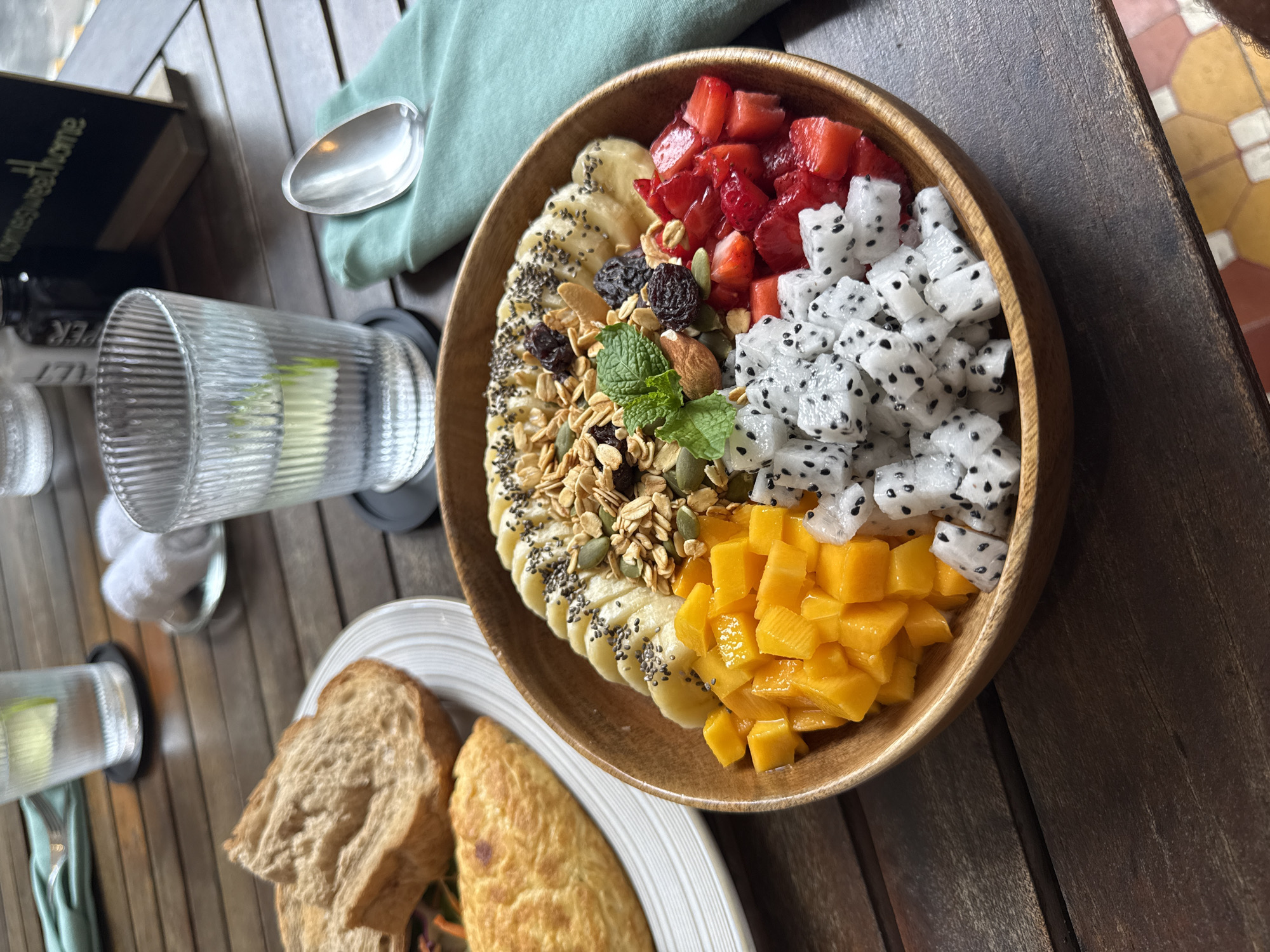 A fruit bowl, containing sliced bananas, strawberries, dragon fruit, and mango, as well as granola, chia seeds, and nuts, sits on a wooden table next to a plate with toast and an omelet. A glass of water with a lime wedge and a spoon on a light green napkin are also present. The image appears to have been taken in a restaurant in Đà Nẵng, Vietnam.