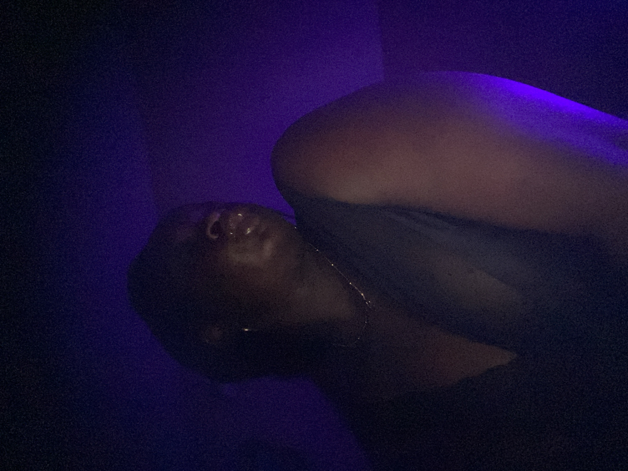 A photograph shows a person with dark skin lying down in what appears to be a dark environment illuminated by purple light. The person's face is visible, positioned at an angle, with a slightly reflective surface suggesting moisture or oil. Part of their upper body, including a shoulder and possibly a chest area, is also visible. A thin necklace is present on the person's neck. The overall scene is low-lit and somewhat obscured. The city or specific location within Maigizo, Nigeria, is not discernible from this image.