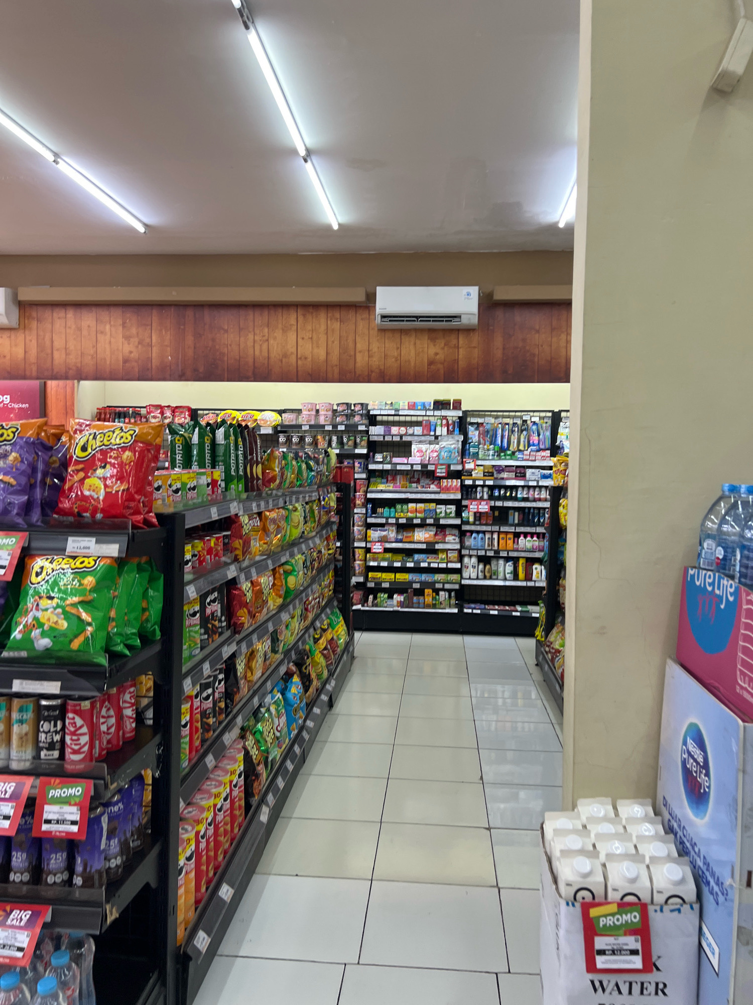 The image shows the interior of a convenience store. The aisle is lined with shelves stocked with various snack food items, primarily chips and biscuits. The shelves are organized with items such as Cheetos, Potato, Kit Kat, and other products in colorful packaging. Further down the aisle, there are shelves stocked with toiletries and personal care products. On the right side of the image, there is a display of bottled water, including Nestlé Pure Life. The store appears to be clean and well-lit with fluorescent lighting. The floor is tiled in a light color. The overall scene is consistent with a modern convenience store in a city like Denpasar, Indonesia, based on the presence of local product packaging and store design common in the region. There are no people visible in the image.