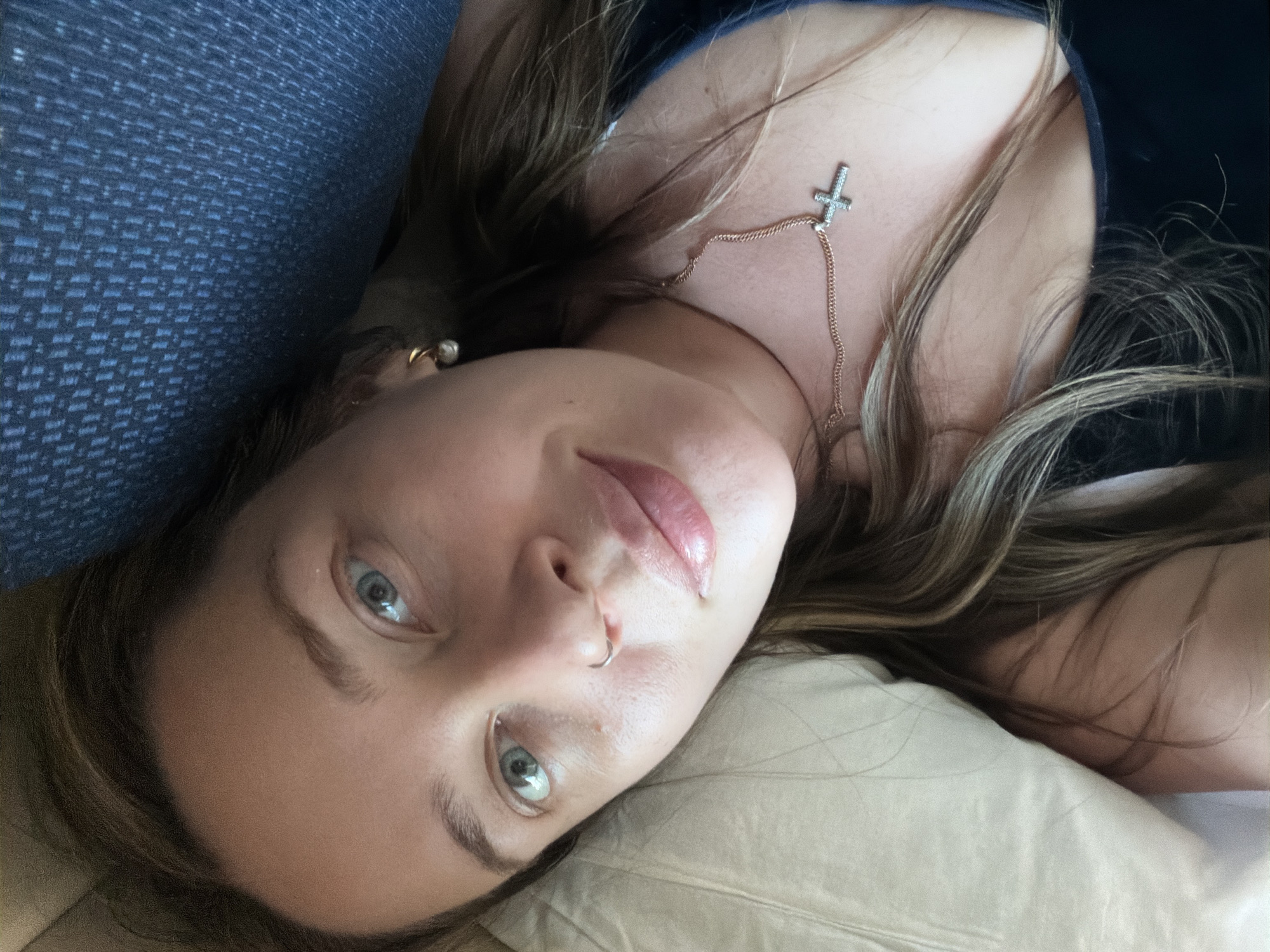 A woman with light brown hair and light eyes is lying down, likely indoors. She is wearing a gold chain necklace with a cross pendant, and a nose ring. She appears to be resting on a light-colored pillow with a blue object behind her head. The image was not taken in Баграш-Бигра, Russia.