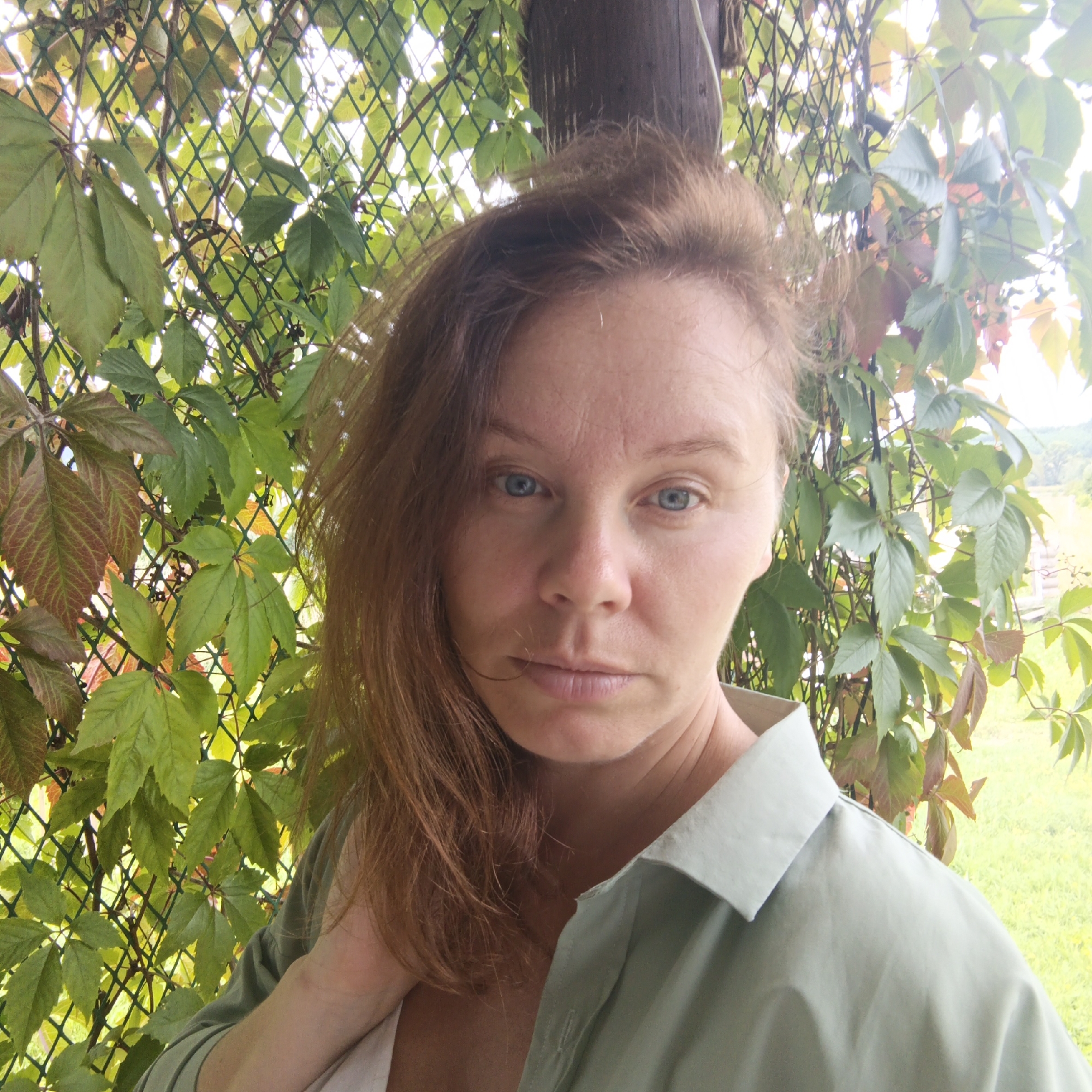 A woman with red hair and blue eyes is looking directly at the camera. She is wearing a light green collared shirt. She is outdoors, with a green wire fence and foliage behind her. The background is slightly blurred, suggesting a shallow depth of field. The lighting appears to be natural daylight. The scene is shot from the chest up. The location is identified as Bolschoj Bukor, Russia.