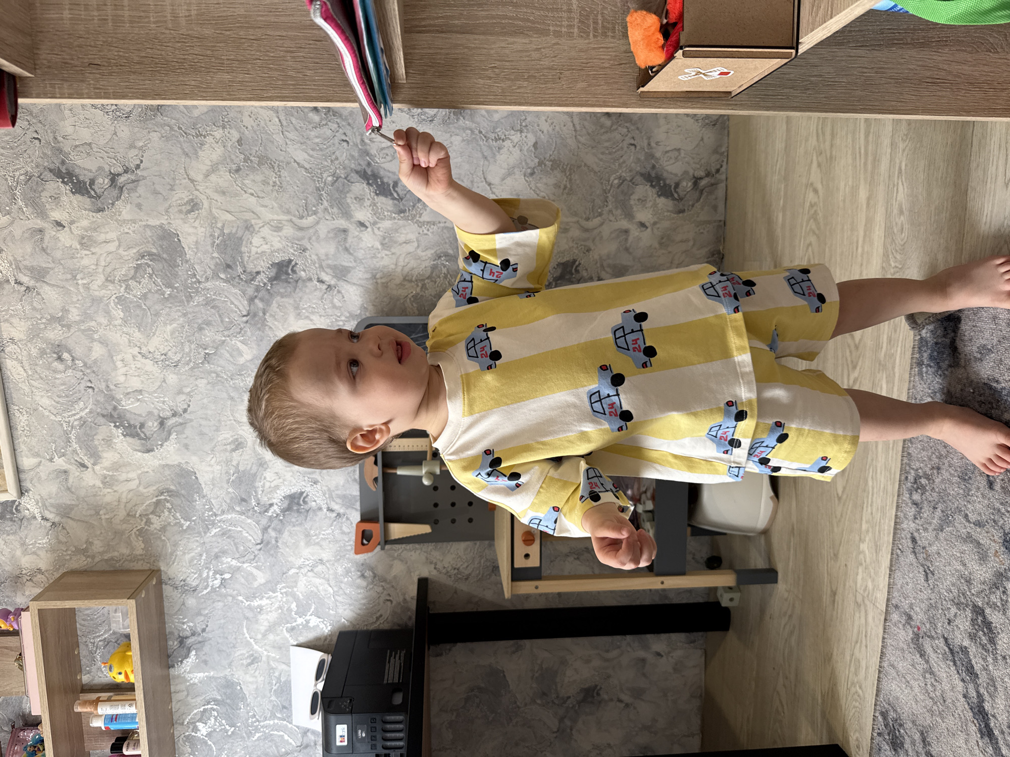 The image depicts a young child standing indoors, wearing a yellow and white striped long-sleeved shirt and matching shorts patterned with blue car-like figures, each displaying the number "24". The child has light brown hair and is looking upwards and slightly to their left with a curious expression. Their right hand is raised, holding a small metallic object.
The setting is an indoor space, likely a child's room or play area. The wall features a grey and white marbled-patterned wallpaper, complementing light wooden-look flooring. A grey patterned rug is visible on the floor.
Various objects are arranged around the child. To their right stands a grey toy workbench equipped with toy tools, including a hammer, saw, and a ruler. Below the workbench, a black printer or similar electronic device is placed, topped with white sunglasses. To the child's left, a wooden shelf unit holds items such as a yellow rubber duck and several bottles. Above the child, colorful fabric items hang from a shelf. An additional wooden shelf holds a small cardboard box containing an orange fluffy toy and an unreadable sticker featuring an arrow. The scene suggests a child engaged in playful exploration in a well-lit home environment.