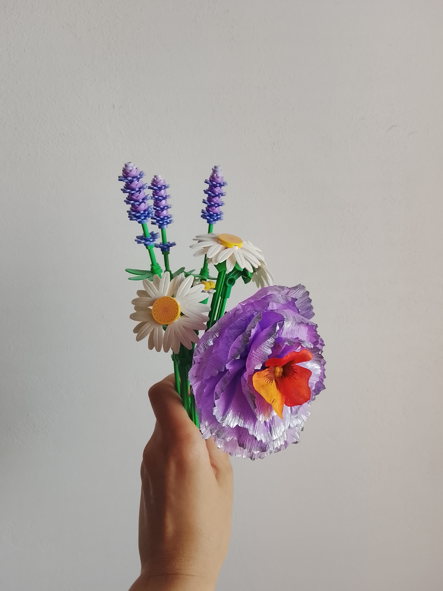 A hand holds a bouquet of artificial flowers, including purple lavender sprigs, white daisies with yellow centers, and a large ruffled purple flower with an orange and red center. The flowers are constructed from what appears to be small plastic bricks, similar to building blocks. The background is a plain, textured white wall. The image is taken from a medium shot, with the hand and flowers filling the bottom right portion of the frame. The lighting suggests an indoor setting with ambient light illuminating the subjects.