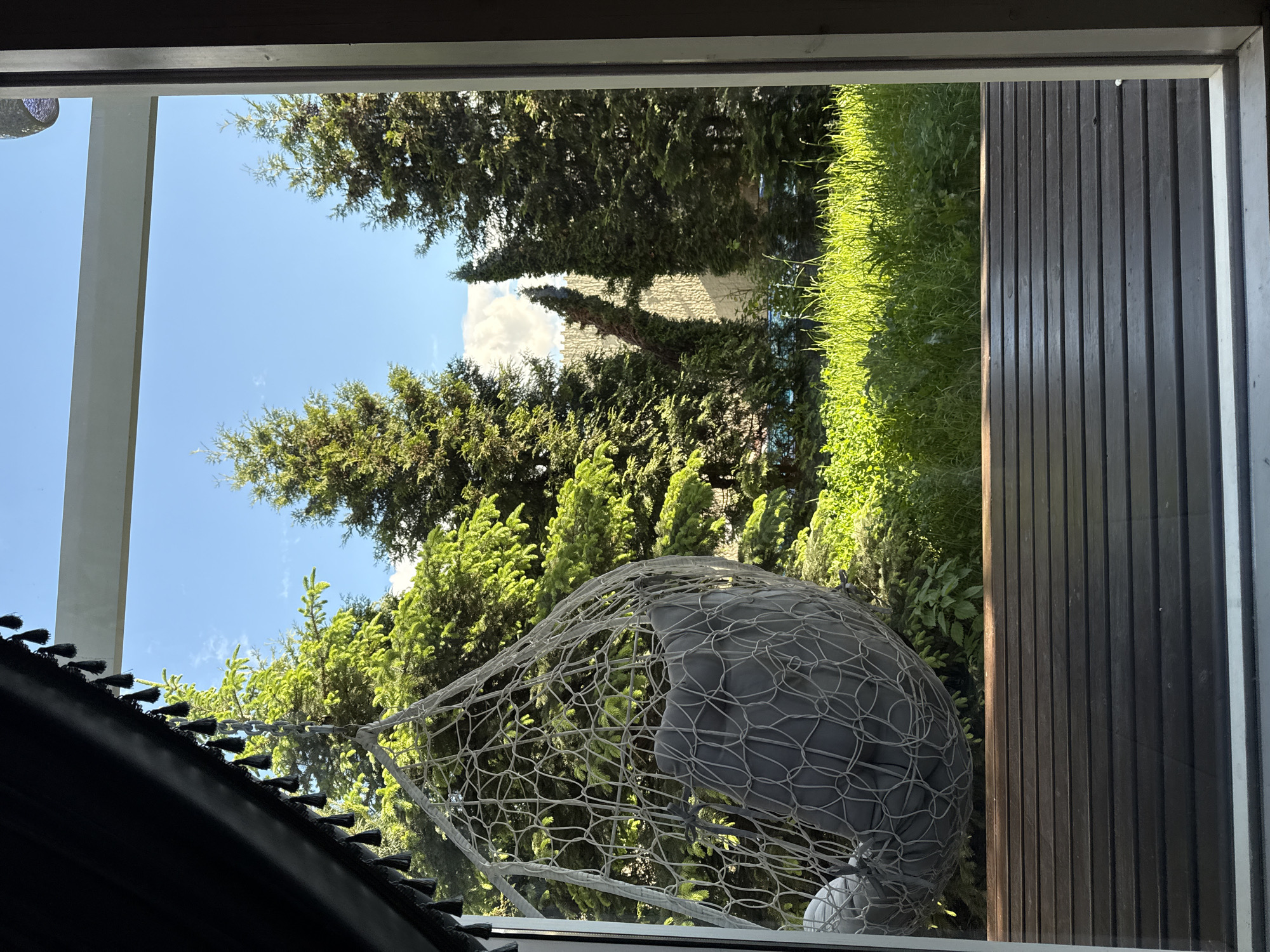 The image shows an outdoor scene viewed through a window. Outside, there are evergreen trees, grass, and a wooden fence. A hanging chair made of netting with gray cushions sits in front of the trees. The sky is blue with some clouds. The window frame is visible, including parts of the interior. The location of Чёрное, Russia, is not discernable from the image.