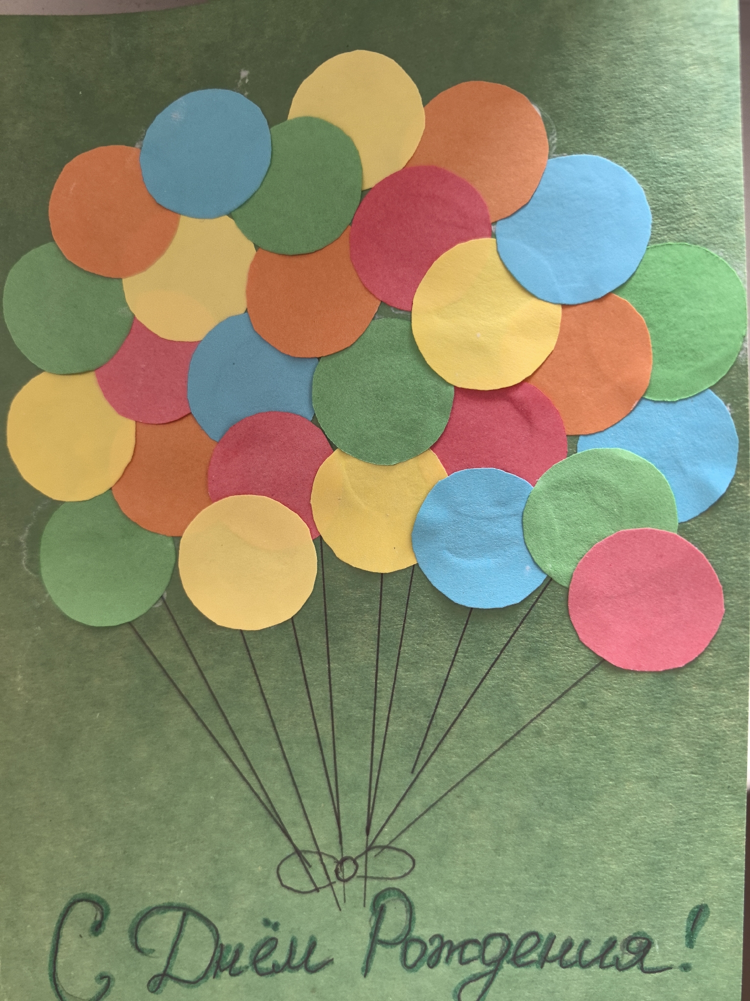 A handmade birthday card on a green background features a cluster of colorful paper circles resembling balloons. Thin black lines represent balloon strings converging at the bottom. Below the balloons, handwritten text in Cyrillic reads "С Днём Рождения!" (Happy Birthday!). The card is lying flat, likely indoors under artificial light. There is no indication of location in the city of Парэчча, Belarus.