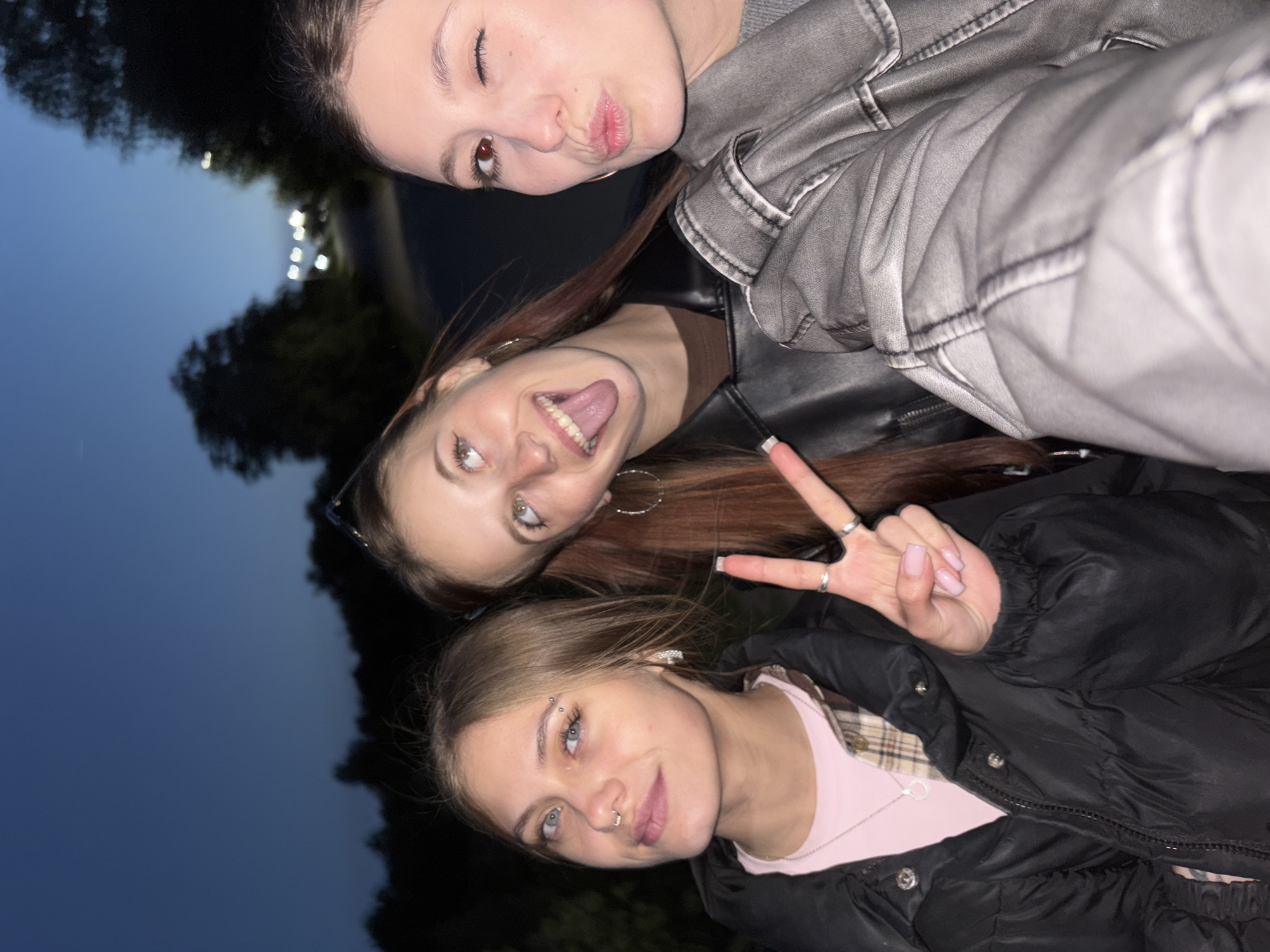 Three young women are posing for a selfie at dusk or night. The woman at the top is wearing a gray leather jacket and winking with pursed lips. The woman in the middle is wearing a black leather jacket and sticking out her tongue. The woman at the bottom is wearing a black puffer jacket over a pink top and plaid shirt and has a septum piercing and an eyebrow piercing. She is making a peace sign with her fingers. The location appears to be outdoors, possibly a park, based on the background which shows trees and dim lighting. The city of Фоки, Russia is not discernible from this image.