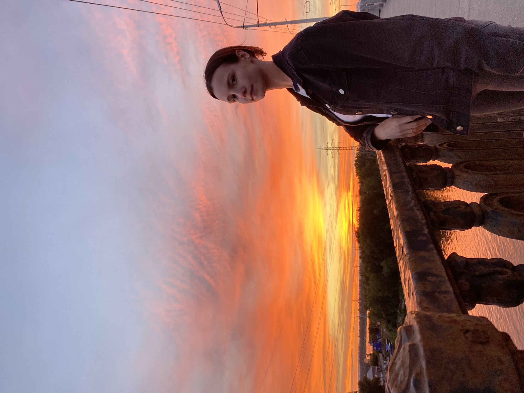 The image captures a young person leaning on a rusty metal railing, gazing out at a vibrant sunset over a body of water. The sky is ablaze with fiery orange and yellow hues, interspersed with streaks of pink and soft lavender clouds, indicating it is dusk. The person is wearing a dark denim jacket over a white t-shirt and has their hair pulled back. A ring is visible on their left hand.
In the background, a tree-lined shore is visible, with some structures and power lines suggesting a somewhat urban or suburban environment. The water below reflects the colors of the sunset, creating a shimmering effect. The overall mood is contemplative and serene, enhanced by the dramatic beauty of the sky. The setting appears to be a bridge or elevated walkway overlooking the water, likely in Novokuybyshevsk, Russia, as suggested by the location context. There is no visible text in the image.