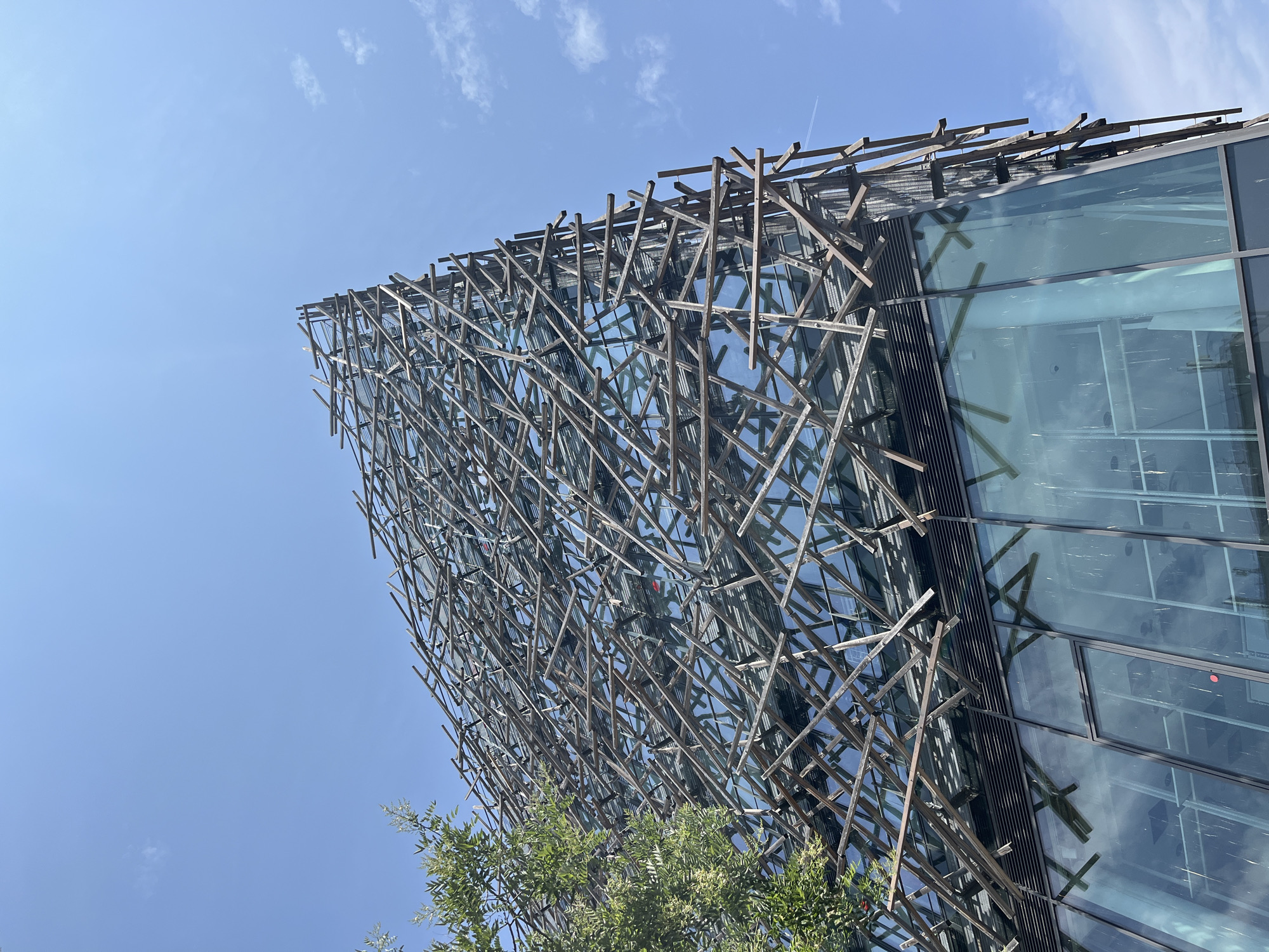 The image displays the exterior of a modern building under a clear blue sky. The building features a distinctive facade composed of a glass curtain wall overlaid with an intricate latticework of thin, dark-colored wooden beams. These beams are arranged in a seemingly chaotic, yet deliberate, pattern, creating a dynamic and textured surface that partially obscures the reflective glass. The glass panels reflect the sky and surrounding environment, adding to the visual complexity.
In the lower left portion of the image, the green foliage of a tree with small white flowers is visible, suggesting an outdoor setting with landscaping. The angle of the shot is looking upwards, emphasizing the height and architectural design of the building. The sunlight suggests it is daytime, and the clear sky indicates fair weather. There are no people or specific activities visible. The overall impression is one of contemporary architecture with a unique, organic-inspired aesthetic. No text is visible in the image.