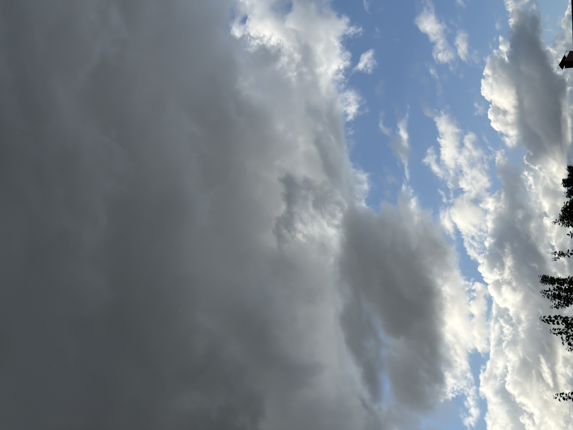 The image depicts a cloudy sky over a rooftop in Узюково, Russia. The sky is a mix of dark, brooding storm clouds in the upper portion and lighter, fluffy white clouds in the lower portion, with patches of blue sky visible. A red brick chimney protrudes from a dark roofline at the bottom right of the frame. Silhouetted tree branches are visible along the bottom edge of the image.