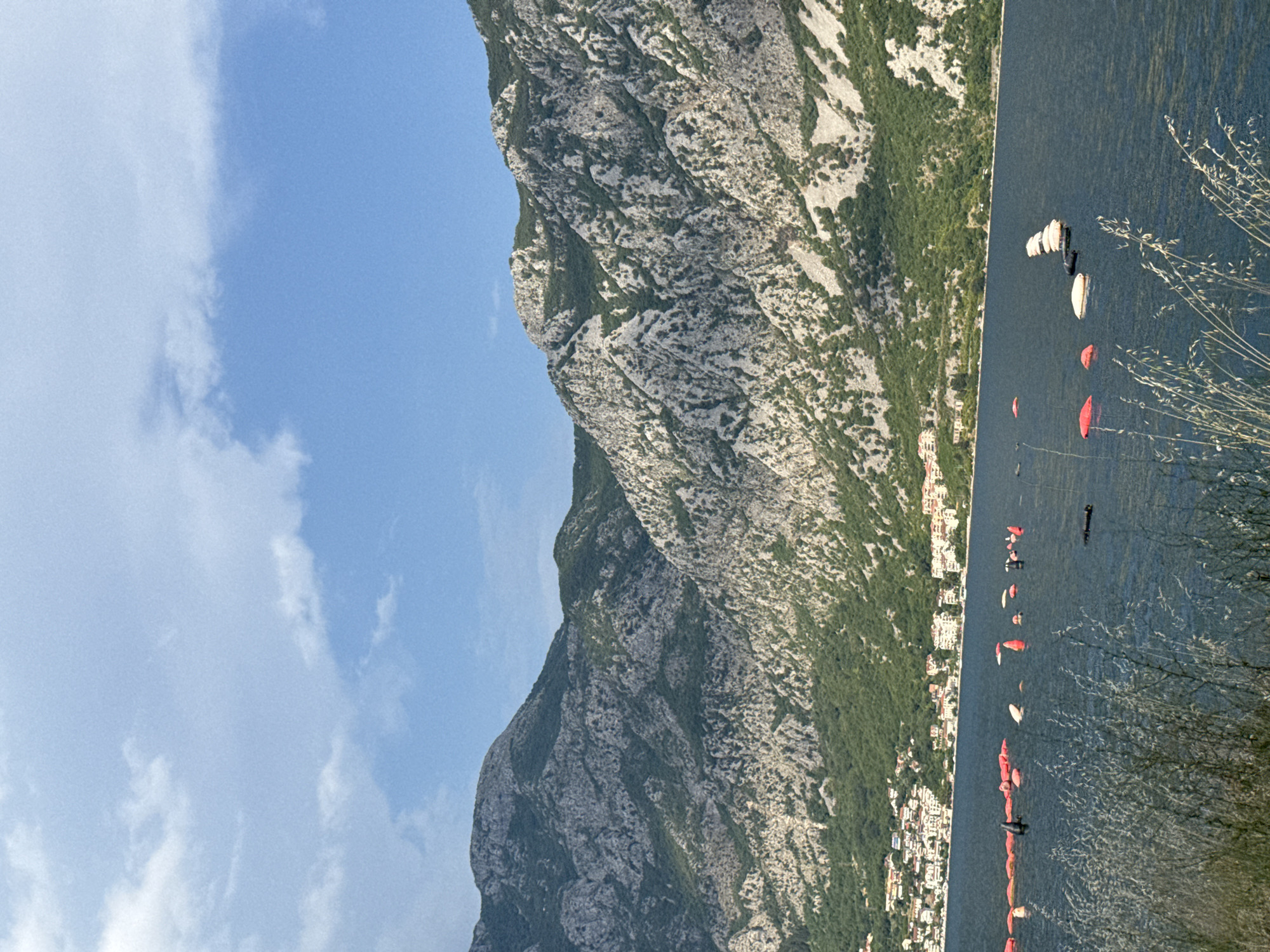 The image depicts a scenic view of the Bay of Kotor, with a focus on the coastline and surrounding mountains. In the foreground, a body of dark water is visible, with a line of red and orange buoys stretching across it. Some of these buoys are spherical, while others are elongated. Further out in the water, there are a few white objects, possibly small boats or buoys. In the mid-ground, a coastal town with numerous buildings is situated at the base of lush green hills. The town features a mix of white and red-roofed structures. Behind the town, imposing, rocky mountains rise steeply, their surfaces textured with grey rock and patches of green vegetation. The sky above is a clear blue with scattered white clouds. The vegetation in the immediate foreground consists of dry, light-colored grasses and other plants. The specific location identified as Strp, Montenegro, is consistent with the visual characteristics of the Bay of Kotor, known for its steep mountains and coastal settlements.
