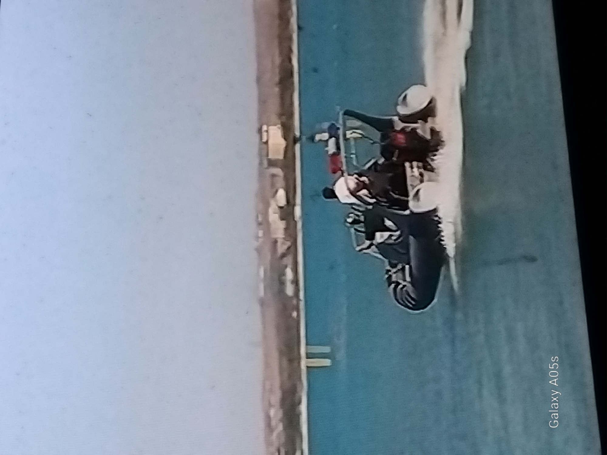 A high-angle, wide shot captures a rigid inflatable boat (RIB) moving across the turquoise water under a clear sky. The boat, a dark color with a rigid hull and inflatable tubes, is occupied by at least three individuals. Two people are visible in the front, with one appearing to wear a white hat and dark clothing, and another further back wearing a dark shirt. A third person, wearing a dark shirt, is visible behind them. Red bags are situated on the boat. The boat is creating a white wake as it moves, indicating speed. In the background, a distant shoreline is visible, lined with buildings and vegetation, suggesting a coastal or lakeside city. Two vertical, light-colored structures are partially submerged in the water near the shoreline. The overall scene depicts a recreational or operational outing on the water.