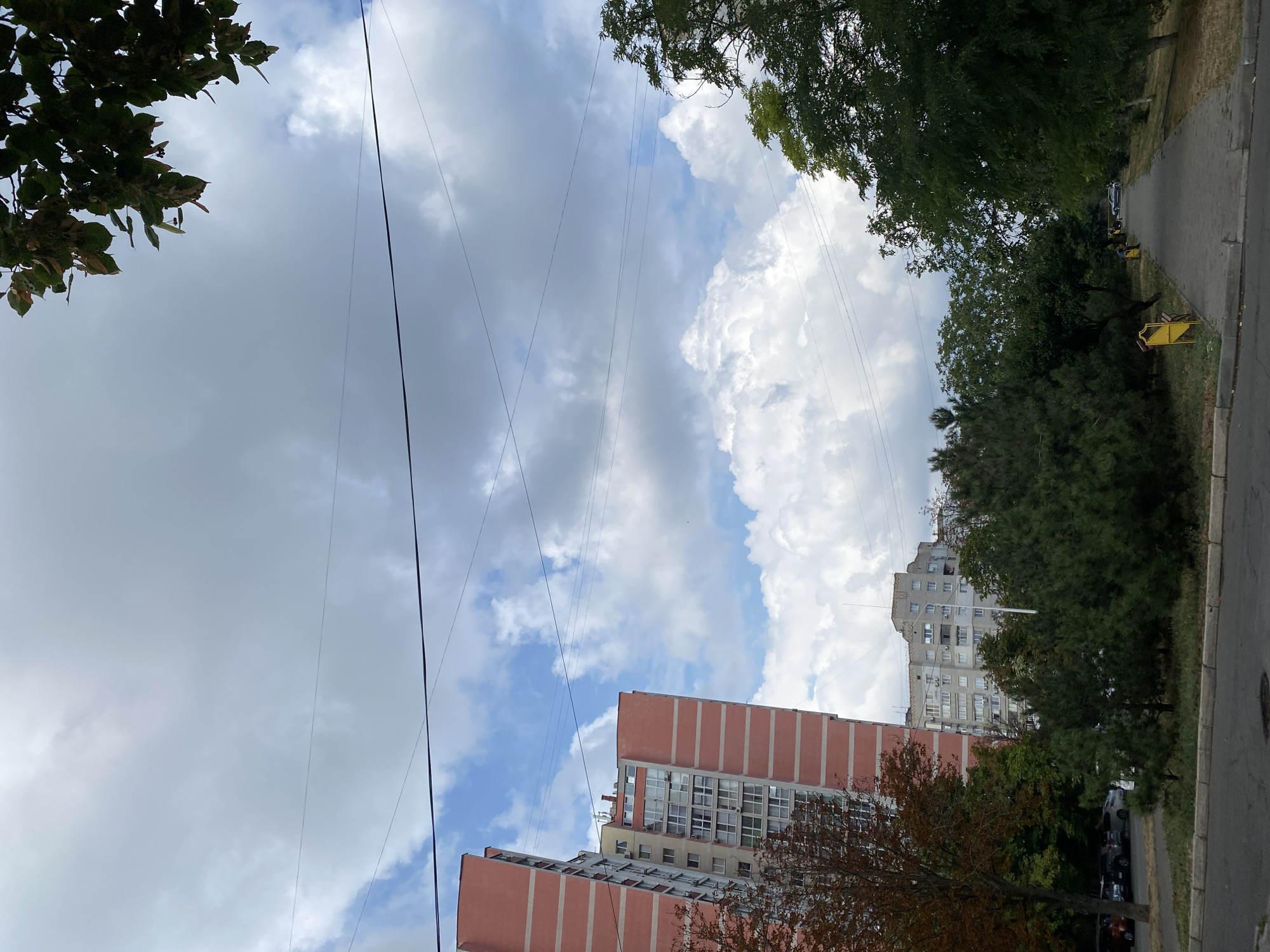 The image shows a street scene in the city of Chornomorsk, Ukraine. On the left, a tall, modern apartment building with pink and beige facade dominates the view. Next to it, a lower, gray apartment building can be seen. In the foreground, a paved walkway runs alongside a strip of green grass and trees. The walkway leads into the distance, where more trees and buildings are visible. Overhead, a cloudy sky with patches of blue fills the upper portion of the image, with power lines crisscrossing the sky. The overall scene depicts a residential area with apartment buildings and green spaces.