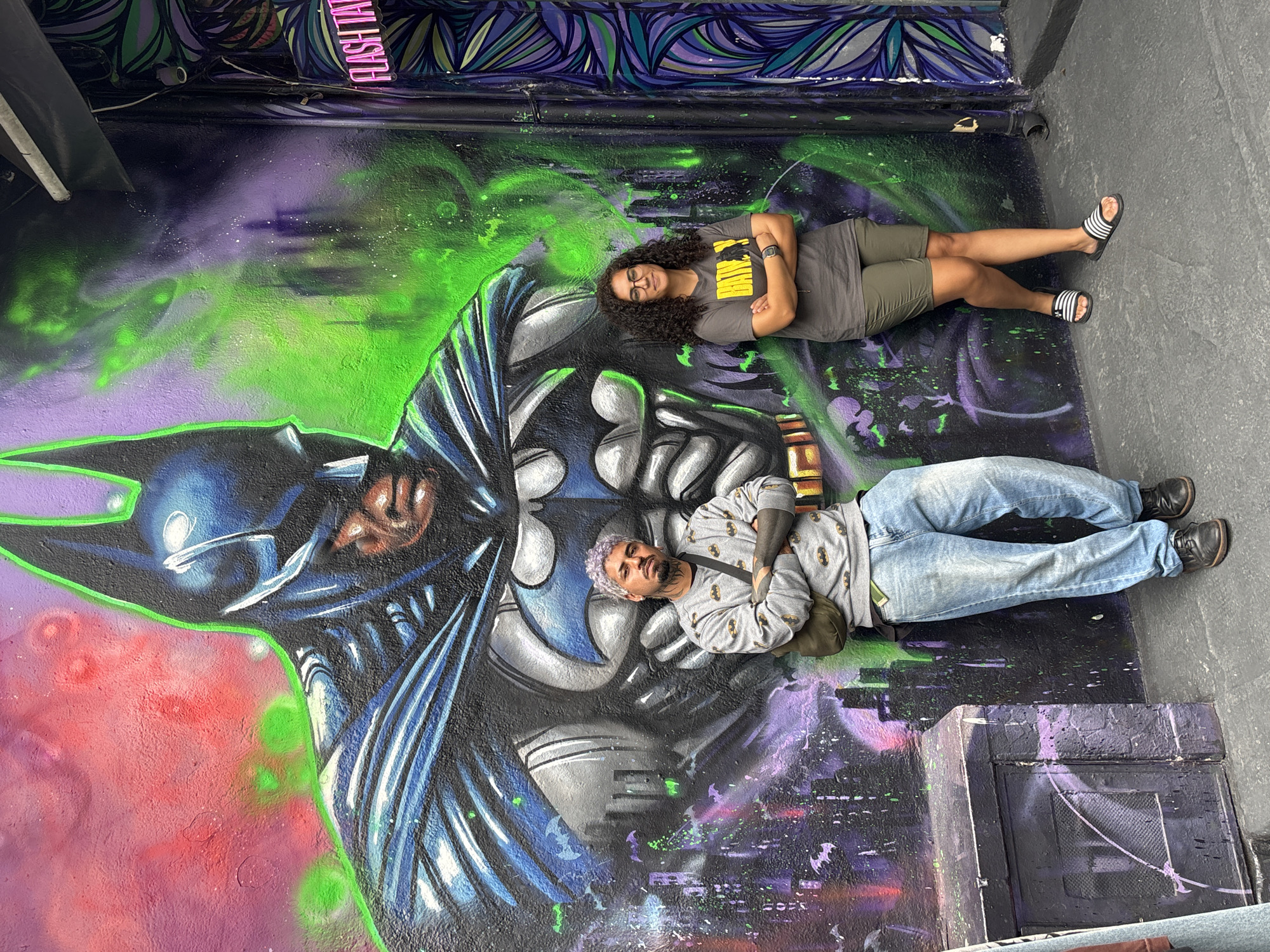 Two people are lying on a mural in what appears to be an alleyway or exterior space. The mural depicts a Batman figure in shades of gray, blue, and black, with splashes of green and purple paint adding to the background. The woman closest to the top of the image is wearing a gray t-shirt with the word "Batman" on it, olive green shorts, and black and white striped sandals. The man is wearing a Batman-themed gray sweatshirt, blue jeans, and black shoes. Both individuals have their arms crossed. Based on the urban graffiti and overall aesthetics, it is suggested that this photograph was taken in São Paulo, Brazil.