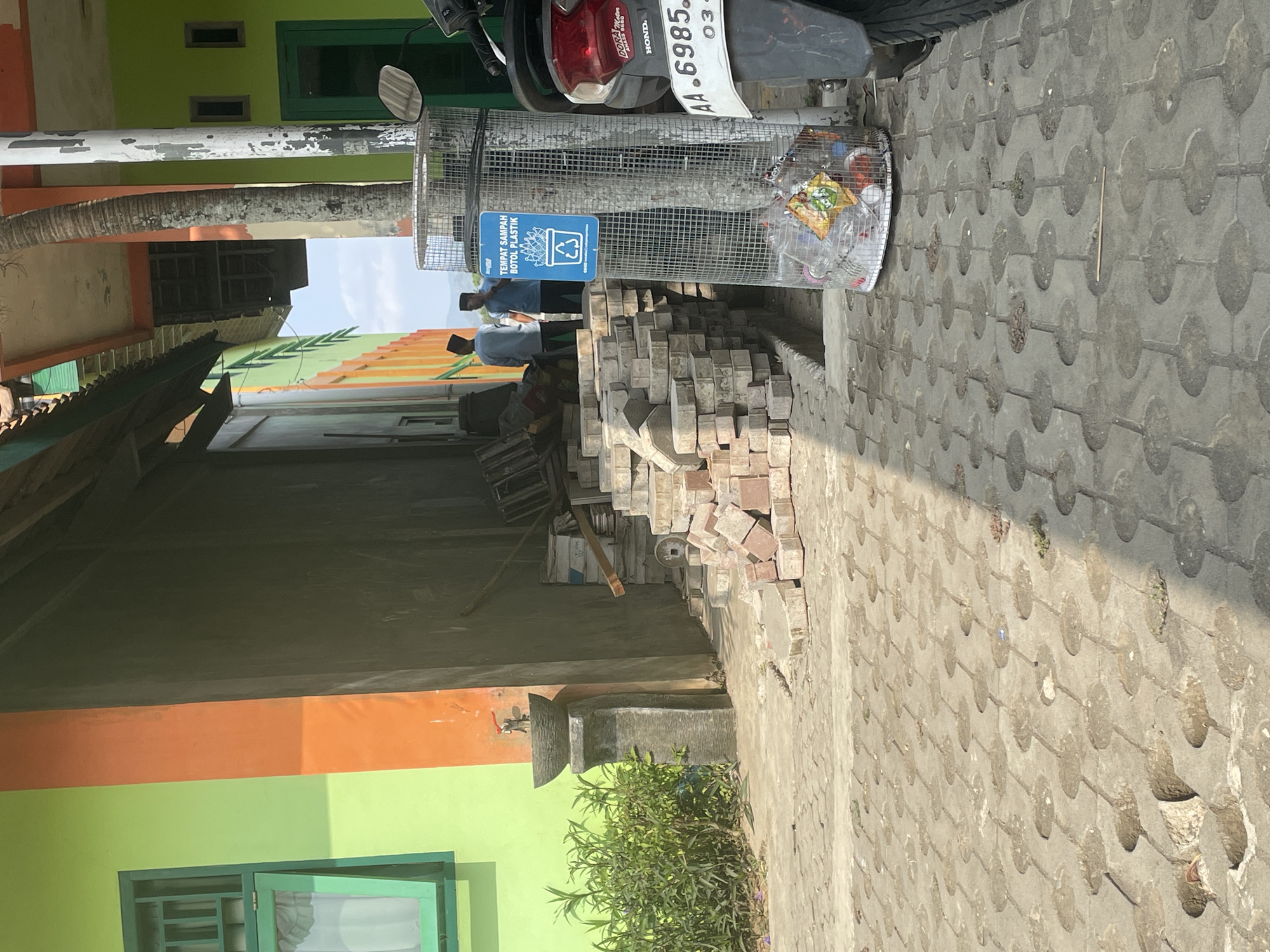 In Borokulon, Indonesia, a scene unfolds with elements of construction, public amenities, and daily life. A vibrant green building with an orange stripe flanks the left side of the image, featuring an open green-framed window with white curtains. Adjacent to it, a concrete pedestal holds a small, dark-colored fountain with a red faucet. In front of the building, a lush green bush grows.
The foreground is paved with interlocking concrete blocks, some of which appear damaged or have circular indentations filled with debris. A pile of rectangular bricks is stacked on the right side of the pathway, indicating ongoing or recent construction. Behind the bricks, a metal trash can, cylindrical and mesh, is visible. A blue sign affixed to the trash can reads "TEMPAT SAMPAH BOTOL PLASTIK," indicating it's a plastic bottle recycling bin. The bin itself contains several clear plastic bottles and colorful wrappers.
Two individuals are present in the mid-ground, standing near the building. Both are wearing dark head coverings. One appears to be wearing a light blue shirt, and the other a darker blue shirt. They are standing near a doorway or opening in the building.
To the right of the trash can, the rear section of a motorcycle is visible, including a red taillight and a white license plate with the inscription "AA 6985 OA". A portion of a palm tree trunk stands between the building and the trash can. The background shows a glimpse of a building with green doors and windows, and distant mountains under a bright sky. The overall environment suggests a warm, sunny day.