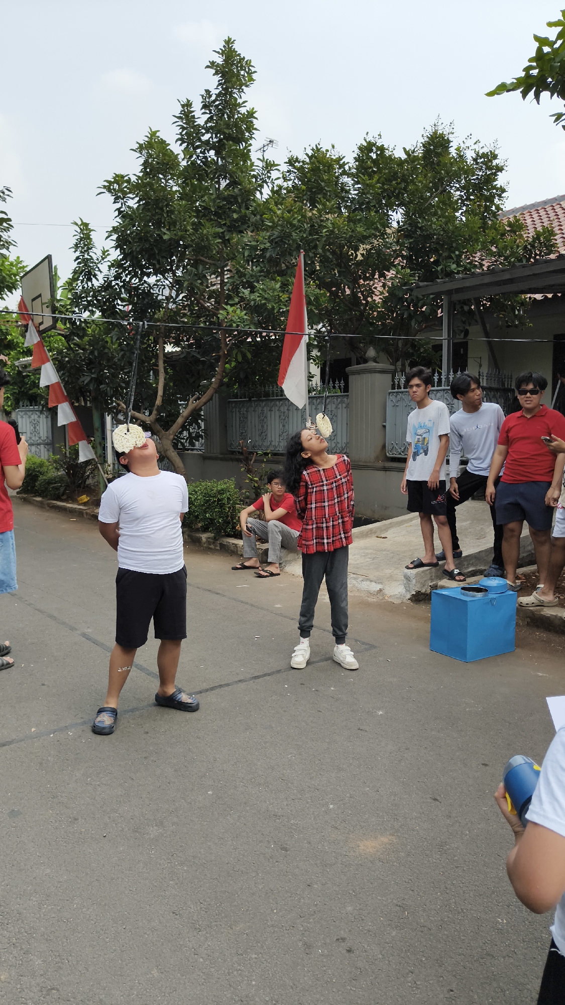 In South Jakarta, Indonesia, a group of children and young adults are participating in a cracker-eating contest. Two children in the foreground are engaged in the activity, with crackers suspended from strings in front of their faces, which they are trying to eat without using their hands. The boy on the left wears a white t-shirt and black shorts, while the girl on the right is in a red and black plaid shirt and grey pants. Several spectators are observing the contest, some standing and others sitting on the curb. The event appears to be taking place outdoors on a residential street, indicated by the presence of houses, trees, and flags, including the Indonesian flag. A blue box is situated on the side of the road, possibly used to collect discarded crackers or for other contest-related purposes. The overall atmosphere suggests a community gathering or a celebration.