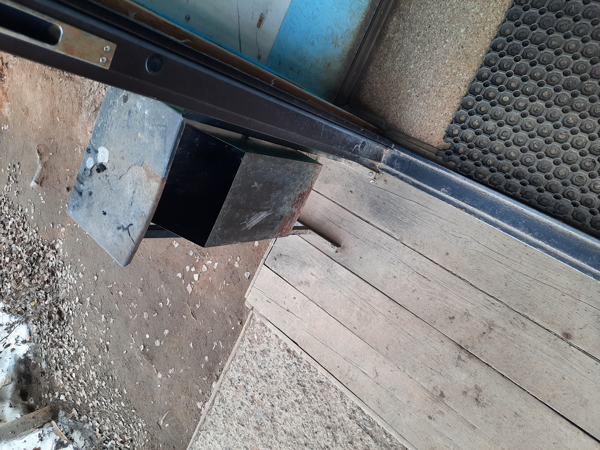 A medium shot, taken from an elevated position, depicts the exterior entrance of a building. A metal trash can, primarily gray and black, sits slightly ajar near the door. To the right of the can, a wooden deck leads to the doorway, bordered by a textured rubber mat and a gray concrete section. The ground visible around the can is littered with leaves and appears partially covered in snow or ice. The frame of the doorway is dark metal, and glimpses of blue and beige are visible through the glass. The image was taken in Krasnogorskoye, Russia.