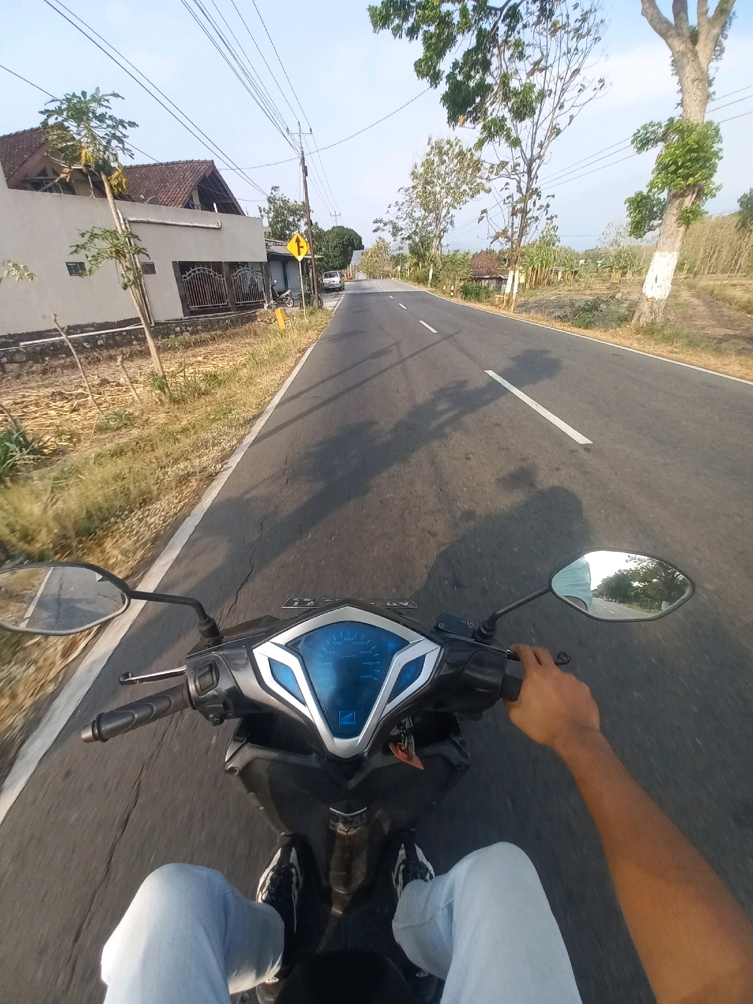 The image captures a first-person perspective of a person riding a motorcycle on a paved road, likely in a rural or suburban area of Baturetno, Indonesia. The rider's hands are visible on the handlebars, and their legs, clad in light blue jeans and sneakers, are positioned in front of the motorcycle's dashboard. The dashboard itself features a digital display with blue illumination.
To the left of the road, a beige house with a brown tiled roof is visible, accompanied by a small tree and a metal fence. Further along the road, a yellow warning sign is mounted on a pole, indicating a turn or hazard. On the right side of the road, there are dry grassy fields, scattered trees, and some sugarcane plants. The road itself has a white line marking its center. The sky is clear and bright, suggesting it is daytime. The overall scene depicts a journey through a landscape characteristic of many Indonesian towns and villages.
