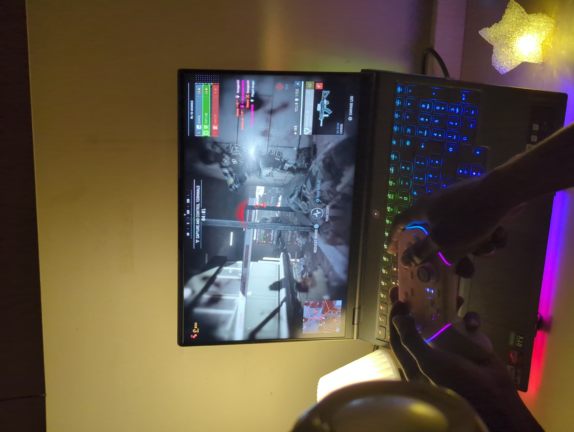 A person is playing a first-person shooter video game on a laptop, using a controller. The game displays capture and control terminals with a timer of 04:01. The laptop has a blue and green backlit keyboard and a visible "RTX" sticker. To the left, the game shows the player's progress in advancing, with "Surge" at 5 and "Fortune" at 6. The game interface also displays player names, distance to enemies (30m), and weapon information (SG 553R CARBINE). Next to the laptop there is a decorative, light-emitting star. The image doesn't contain sufficient information to confirm a location in Jiangjin, China.