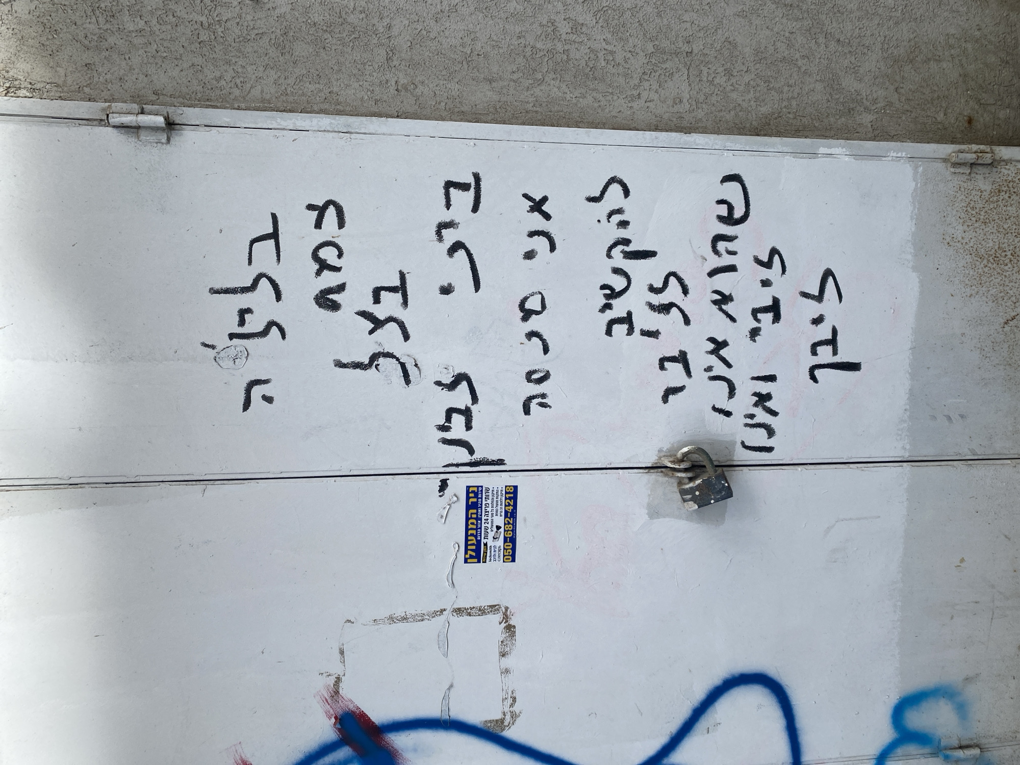 The image displays a weathered, off-white metal double door or gate, secured by a dark metal padlock positioned centrally between the two panels. The surface exhibits significant wear, with peeling paint, grime, and several layers of graffiti.
On the left door panel, fragmented blue spray paint forms abstract shapes, with remnants of red spray paint visible beneath. A rectangular blue and white sticker, advertising "ניר המנעולן" (Nir the Locksmith) with services like "שירות מהיר ואמין" (fast and reliable service) and "שירותי פריצה 24 שעות" (24-hour emergency services), along with the Israeli phone number 050-682-4218 and website nirthelocksmith.co.il, is affixed to the upper left portion.
The right door panel is covered with multiple lines of black Hebrew script, applied with what appears to be a marker or paint. The text includes phrases such as "בלביה" (in her heart), "צמח בצל" (grows in the shade), "ביתי לבן" (my home is white), "אני ססמה" (I am a password), "להקשיב לצבי" (to listen to Tzvi), "שהוא אינו" (that he is not), and "צבי ואנו לצבי" (Tzvi and we to Tzvi). Some of these lines have faint red marks interspersed with the black.
The right edge of the right door panel features two visible metal hinges, connecting it to an adjacent wall. This wall is textured, appearing as a light, coarse stucco or rendered surface, consistent with urban architecture. The overall scene suggests a back entrance or utility access in an urban environment, specifically located in Tel-Aviv, Israel, as indicated by the Hebrew text and phone number. No people or animals are present.