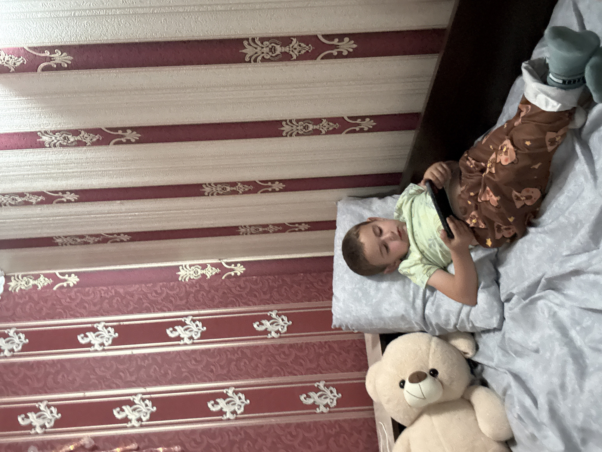 A young boy with short brown hair, wearing a light green t-shirt and brown pajama pants with a bear pattern, is lying on a bed and playing a video game on a dark-colored handheld device. He is propped up against a light blue pillow, and a large, light beige stuffed teddy bear is positioned to his left. The bed is covered with a light gray sheet. Behind the bed, the wall is covered in striped wallpaper with alternating vertical bands of dark red with a floral pattern and cream-colored textured stripes. The scene appears to be indoors, likely a bedroom, in Svoge, Bulgaria.