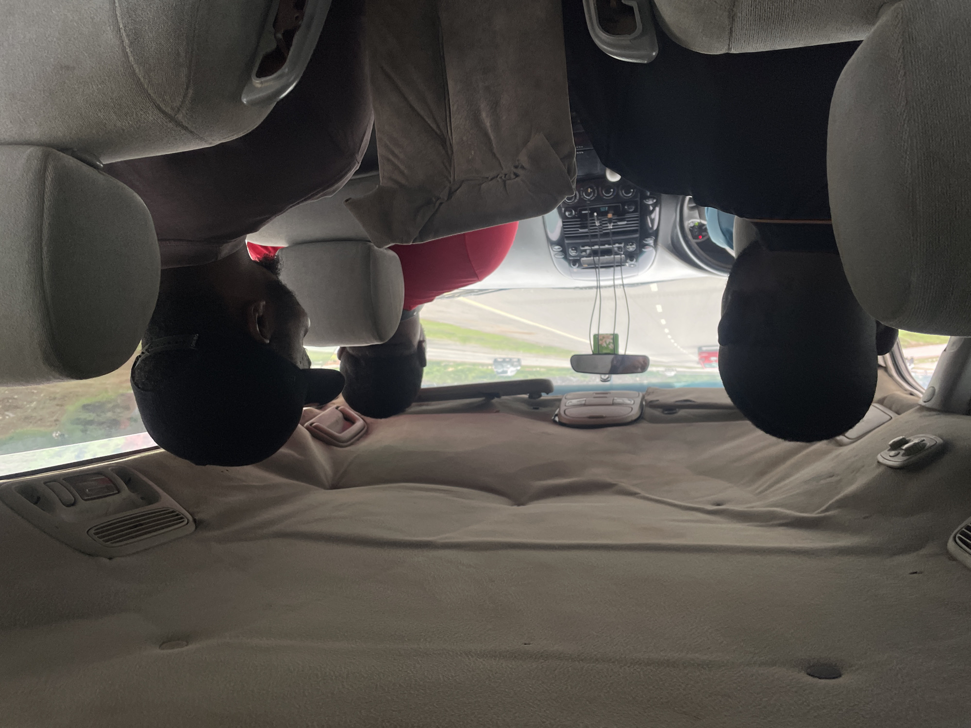 In Kugbo, Nigeria, four individuals are captured from behind inside a vehicle traveling on a highway. The perspective is from the rear passenger seats, looking forward. A man in a black shirt is seated in the left rear, facing forward. To his right, another man in a red t-shirt is visible, likely the front passenger or driver. In the right rear seat, a man wearing a black baseball cap is seated. The vehicle's interior shows worn fabric on the ceiling and seats. The highway stretches ahead, with other vehicles in the distance. The scene depicts a journey through Nigeria, highlighting the interior of a car in transit.