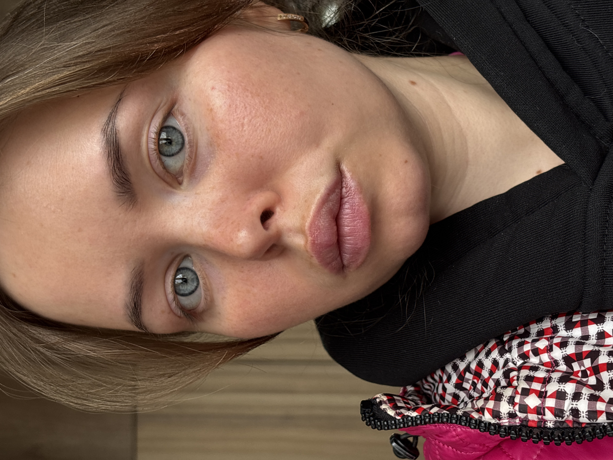The image shows a close-up of a woman's face. She has light skin, freckles, and light blue eyes. Her hair is blonde/light brown. She is wearing a black garment, and part of a pink and patterned jacket is visible. The image is rotated. The location is not discernible from the image.