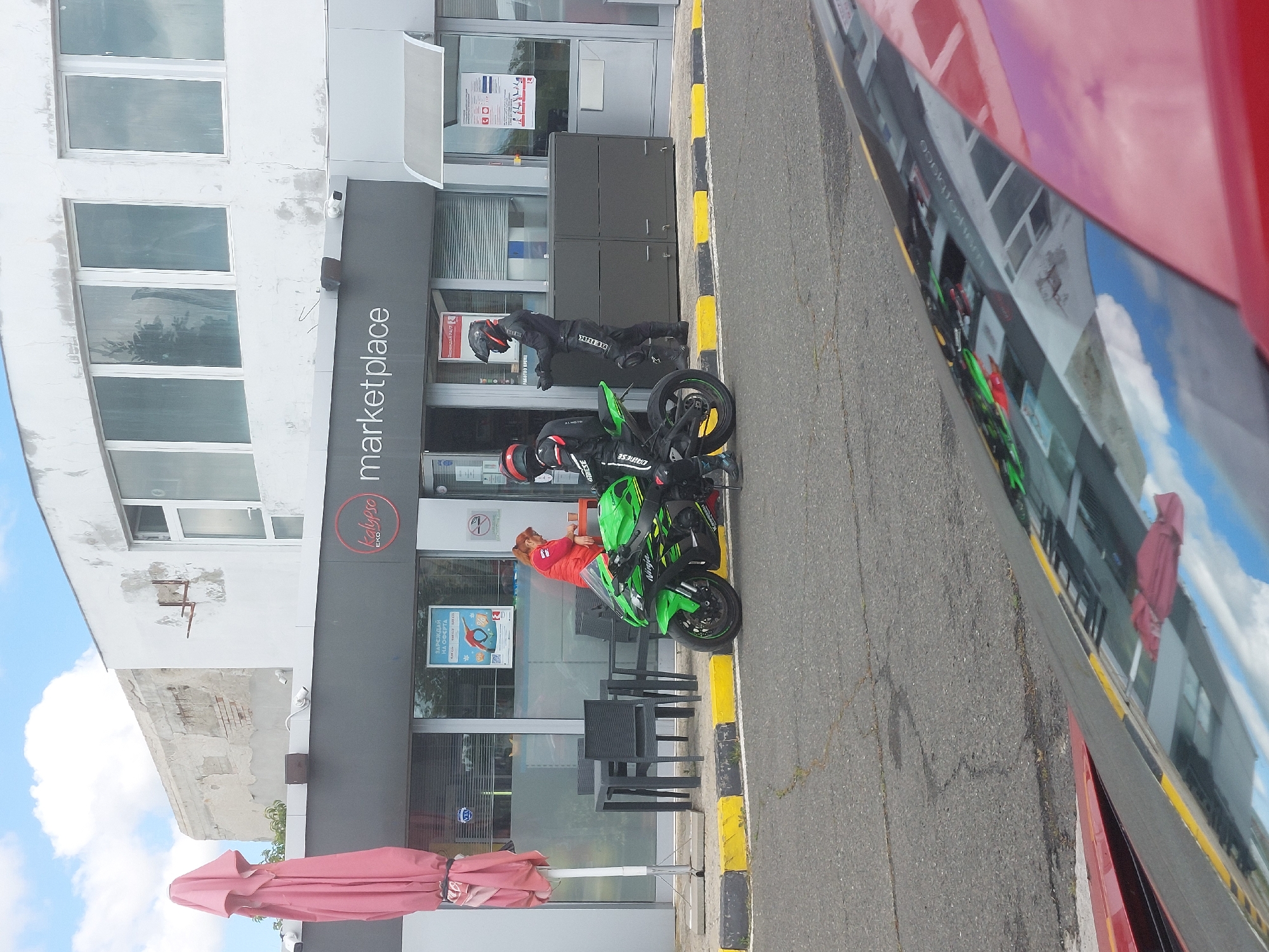 The image depicts the exterior of a building with the signage "kalypso marketplace eko". Two motorcycles are parked in front of the building, alongside the curb with black and yellow stripes. One person in a red jacket is sitting outside the store. Another individual is seen in motorcycle gear beside one of the motorcycles, and a second person also in motorcycle gear is near the other motorcycle. A red parasol is visible, and chairs and tables are set up on the pavement. The reflection of the building is seen in a red car. The scene is likely in the city of Aheloy, Bulgaria, based on the branding on the building.