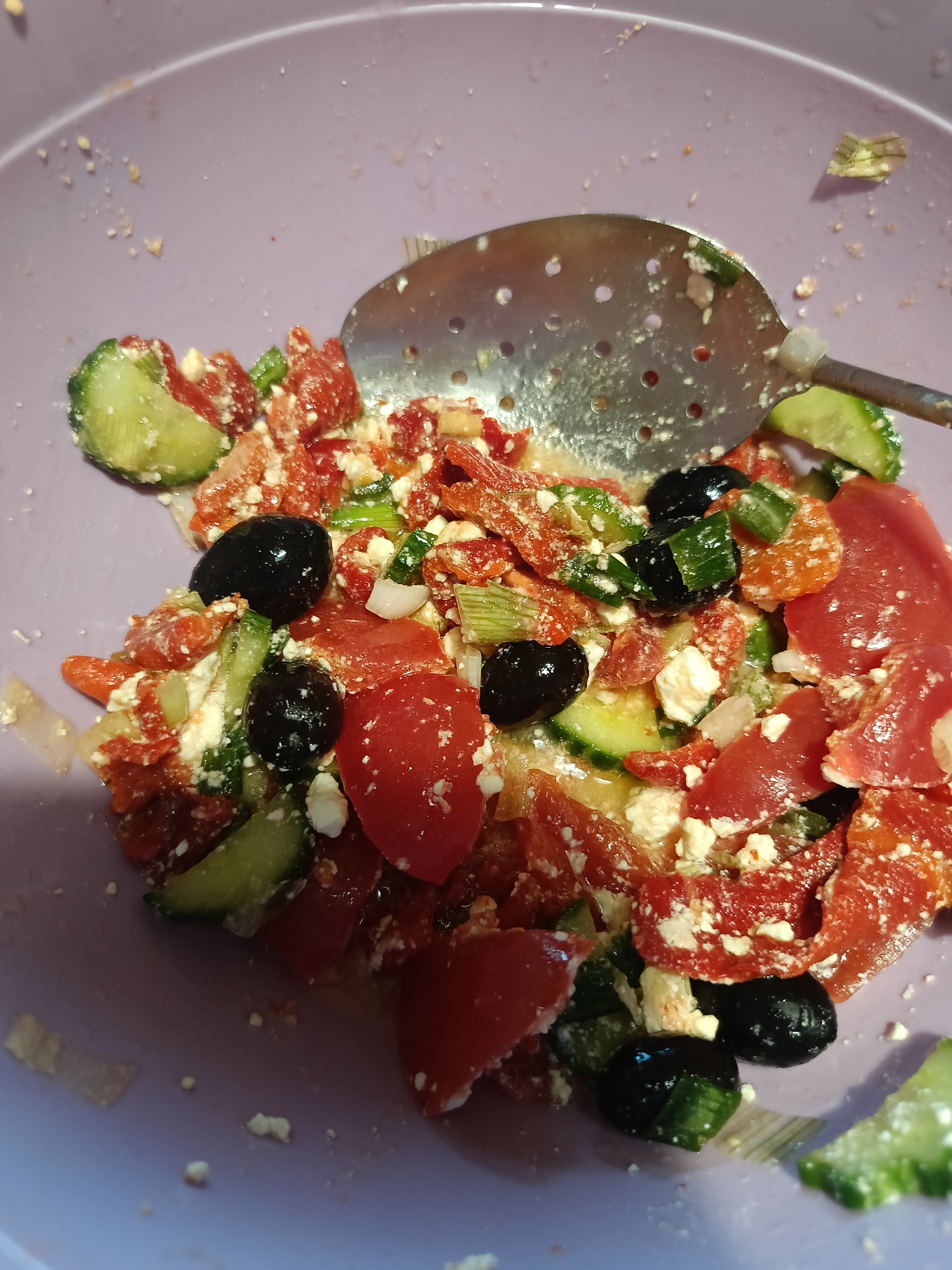 In Varna, Bulgaria, a close-up, high-angle shot captures a portion of a purple bowl filled with a chopped salad. The salad consists of diced tomatoes, sliced cucumbers, black olives, crumbled feta cheese, and chopped green onions. A metal slotted spoon is positioned in the upper right quadrant of the bowl, resting partially on the salad. The bowl itself appears slightly dirty, with scattered crumbs and a few droplets of liquid on the interior rim. The lighting is overhead, creating shadows within the salad. No people are visible in the image, and the background is out of focus.