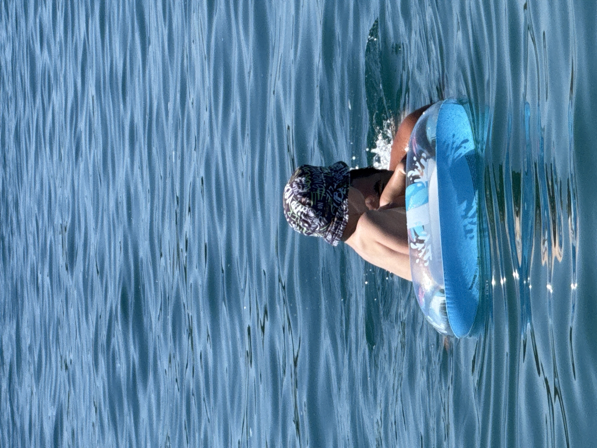 The image displays two individuals partially submerged in tranquil blue water under natural daylight in Boreti, Montenegro. A person, appearing to be a child, is positioned in the foreground, facing away from the camera, wearing a dark-colored bucket hat with white and light green abstract patterns. A blue and white inflatable swim ring is visible around their torso. Behind and slightly to the right of this individual, a second person is partially discernible, with dark hair, a portion of their back, and an arm visible above the water line, seemingly supporting the first individual. Splashes of water are present around the second person. The water surface exhibits gentle ripples, reflecting ambient light. No shoreline or other land features are visible in the background.