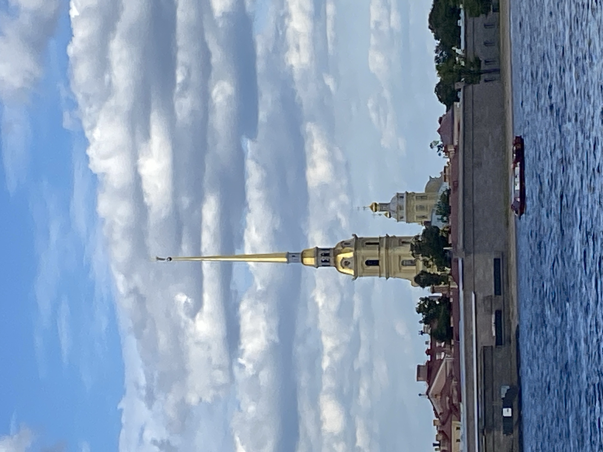 The image depicts the Peter and Paul Fortress, a historical citadel located on Zayachy Island in Saint Petersburg, Russia. The most prominent feature is the spire of the Peter and Paul Cathedral, which is golden and topped with an angel. The cathedral itself is a light yellow color with arched windows. Adjacent to the cathedral is another building with a smaller golden dome. These structures are situated behind a thick, grey stone wall that forms the fortress's perimeter.
In the foreground, a body of blue water, likely the Neva River, stretches across the bottom of the frame. A red boat with a dark roof is moving on the water. The sky above is a mix of blue sky and white, fluffy clouds. The overall impression is a bright, somewhat overcast day. The architecture suggests a historical European setting.
