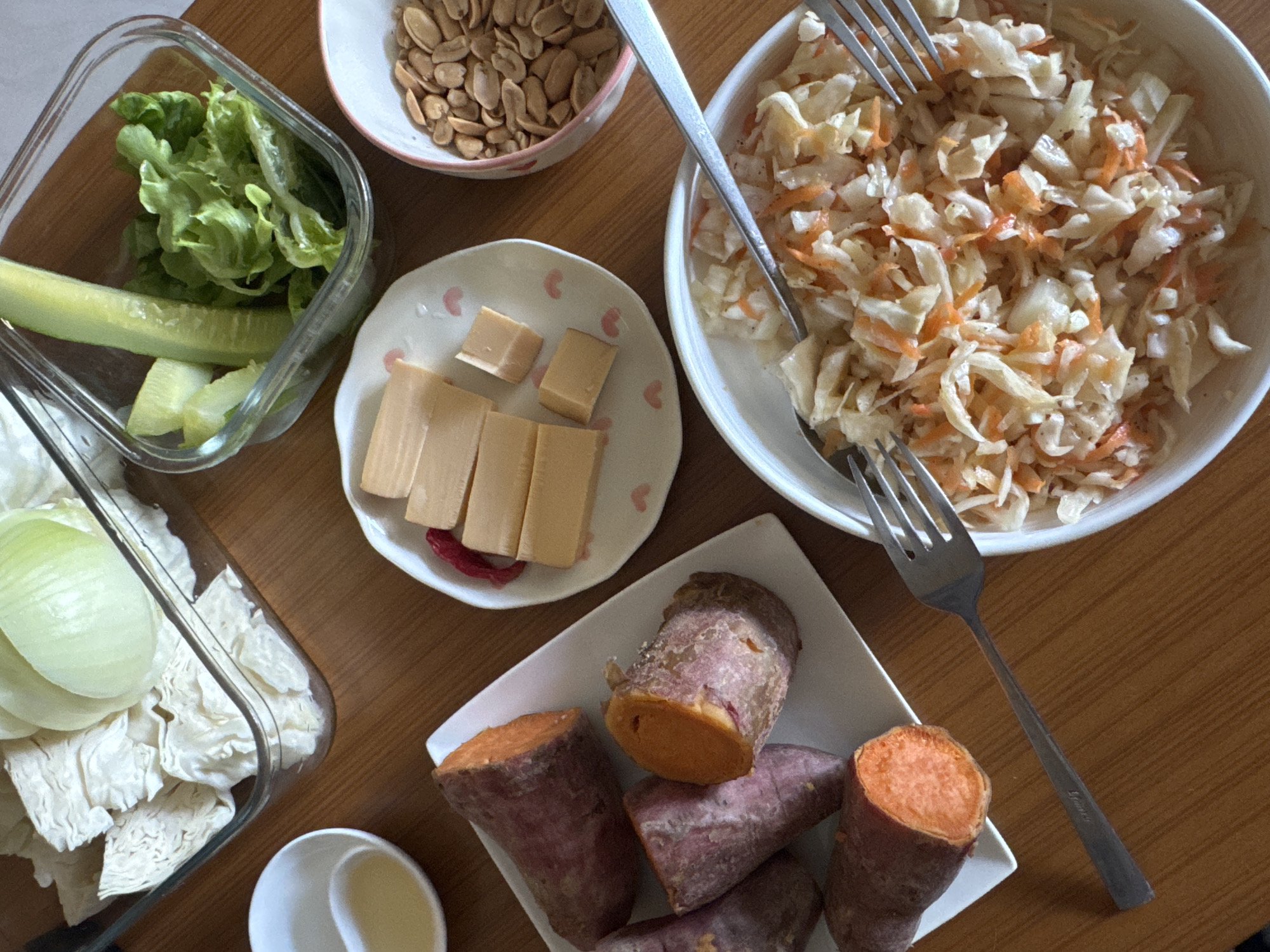 The image displays a meal spread arranged on a light brown wooden table, likely indoors. No specific identifying features are present to determine the geographic location, including the city of Baan Nok Sai, Thailand.
The table holds multiple items presented in various containers. In the upper left, a clear rectangular glass dish contains chopped white cabbage leaves and a half-cut pale green onion. Adjacent to it, a small white bowl holds a pale yellow liquid.
In the upper right, a clear square glass dish contains slices of green cucumber and fresh green leaf lettuce. Next to it, a small white bowl with a pink-patterned rim is filled with shelled roasted peanuts.
A white rectangular plate on the left side holds several cooked sweet potatoes, some whole and some halved, revealing their bright orange interiors.
On a smaller white plate with pink heart motifs, several light brown rectangular food items are arranged, some whole blocks and some cut into smaller cubes. A few small red pieces are visible near them.
In the lower right, a white circular bowl contains a prepared salad resembling coleslaw, consisting of shredded white cabbage and grated orange carrots. Two silver-colored metal forks rest across and near this bowl. The overall scene suggests a prepared meal or ingredients ready for consumption.