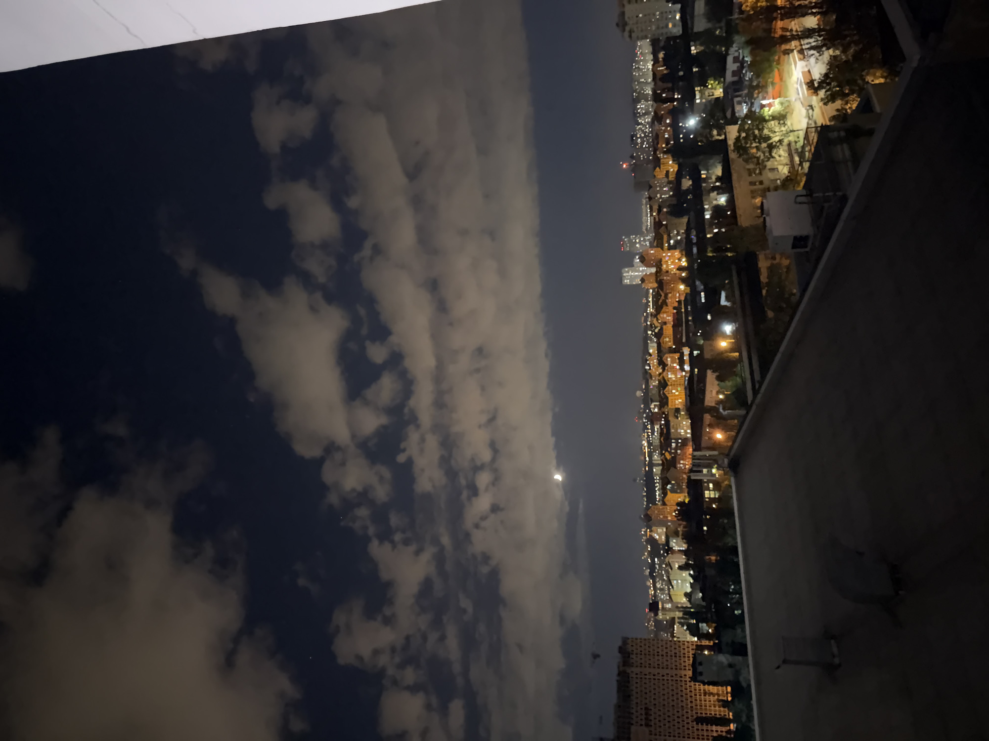 An aerial nighttime view of a city skyline, presumably Kyiv, Ukraine, captured from a rooftop. The foreground is dominated by the flat, dark surface of the rooftop, with visible ventilation shafts. In the mid-ground, a dense urban landscape unfolds with buildings illuminated by various lights, indicating a residential or commercial area. Some taller structures stand out against the horizon. The background features a dark sky with scattered clouds that are partially lit from below by the city lights. A crescent moon is visible in the sky. The overall scene depicts a nocturnal cityscape with a sense of depth and scale.