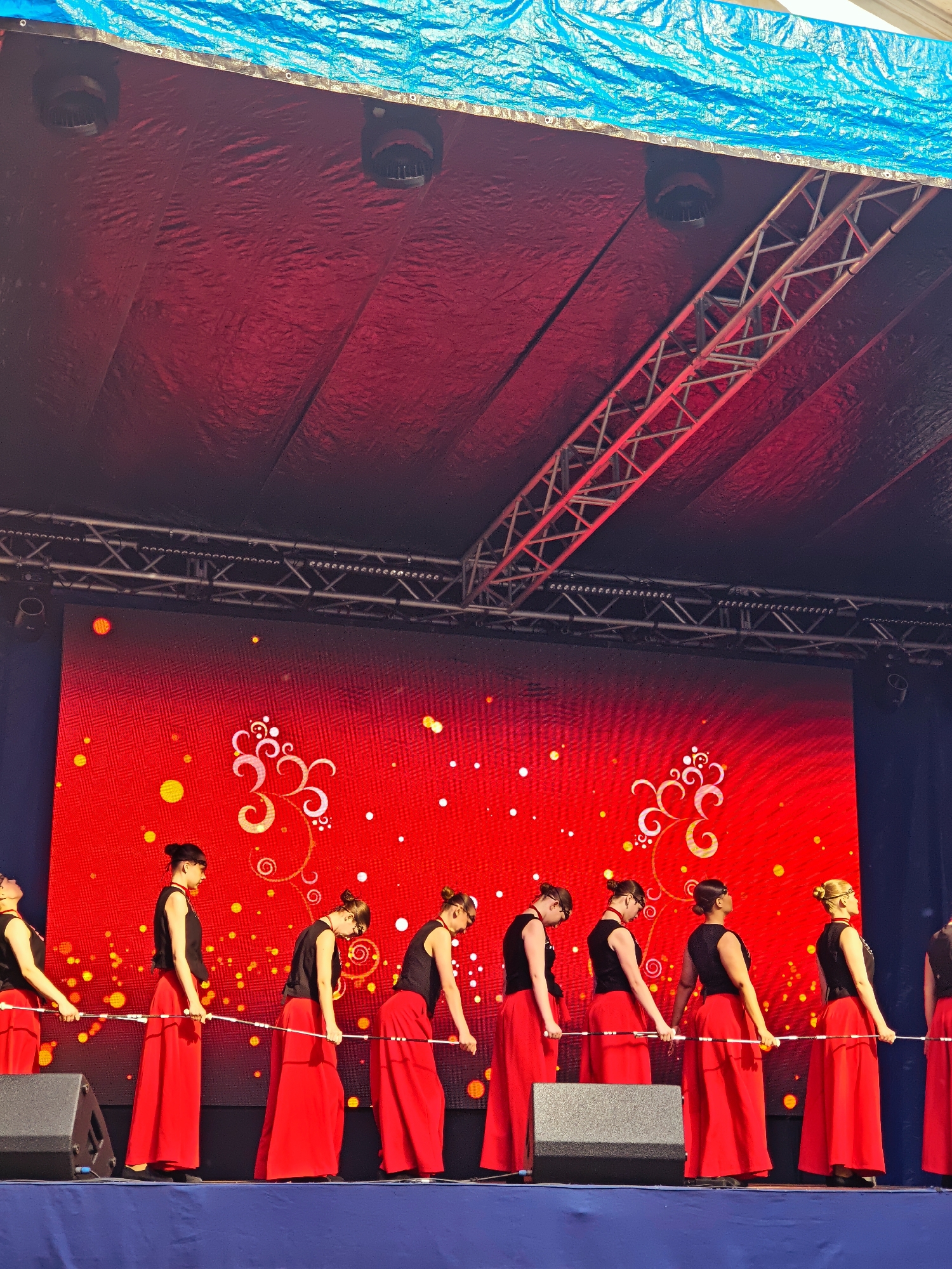 A group of dancers in red skirts and black tops are performing on a stage in Kimry, Russia. They are holding a white pole and moving in unison. The stage backdrop is a large red screen with a pattern of white swirls and yellow dots. The stage is lit with red lights, and there is a blue tarp overhead. The dancers are all young women, and they are all looking down at the pole. There are several speakers on the stage, and a truss system is visible above the screen.