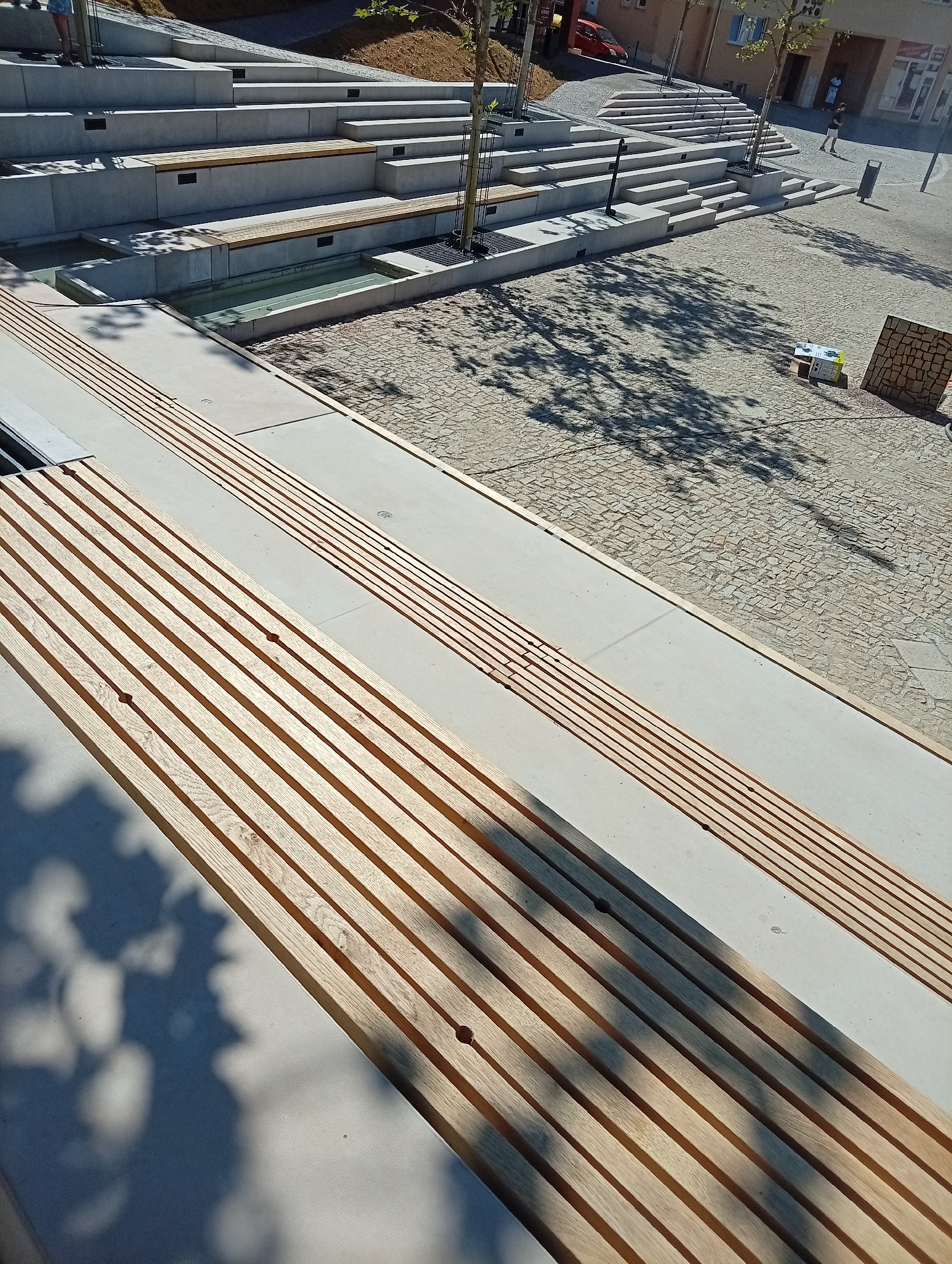 This image depicts a newly constructed public seating area with tiered concrete steps and wooden benches, possibly located in Tišnov, Czechia. In the foreground, several rows of slatted wooden benches are visible, some casting shadows on the light-colored concrete. Behind these benches, a tiered seating structure made of concrete forms multiple levels, some of which also incorporate wooden bench seating. A shallow water feature or channel is present between two of the concrete tiers. The ground surface is paved with small cobblestones, and trees with casting shadows are scattered throughout the scene. In the background, a red car is parked near a building, and a few people are visible, one walking on the paved area. The overall impression is of a modern, open-air public space designed for relaxation and gathering.