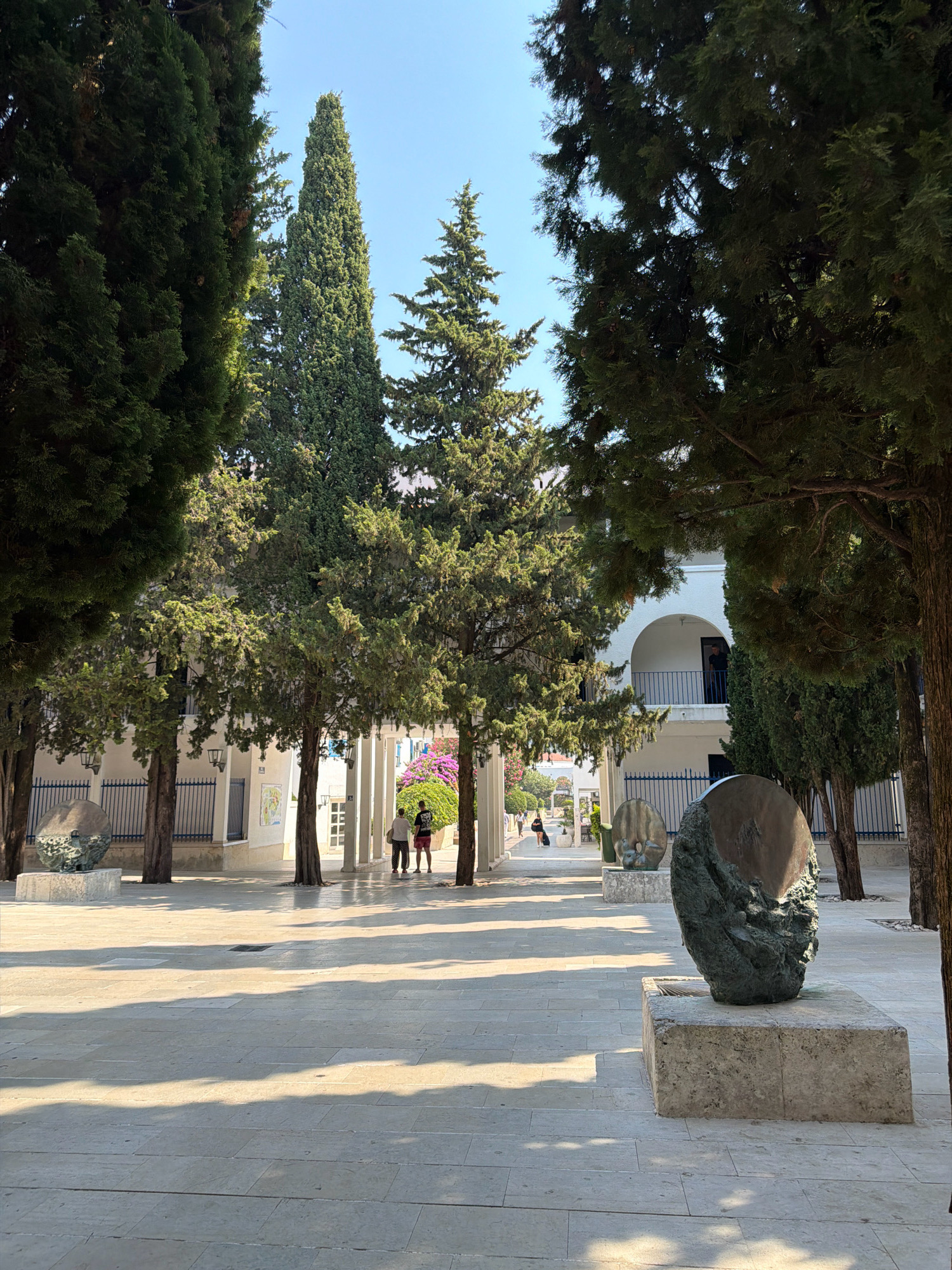 The image captures a sunlit courtyard with paved stone flooring, featuring several prominent cypress trees. In the foreground, a large, abstract sculpture made of dark metal rests on a stone pedestal. To the left, another similar sculpture is partially visible.
In the middle ground, two individuals, one wearing a white shirt and the other a red shirt, stand near a colonnaded building. Further back, through an archway, more people can be seen walking and a vibrant purple bougainvillea plant adds a splash of color. The architecture appears to be white with blue railings, characteristic of Mediterranean coastal towns.
The scene is identifiable as the courtyard of the Saint Ivan Church in the Old Town of Budva, Montenegro, given the distinctive architecture and the presence of the sculptures, which are part of the local art installations. The time of day appears to be late morning or early afternoon, judging by the long shadows cast on the courtyard. The sky is clear and blue.