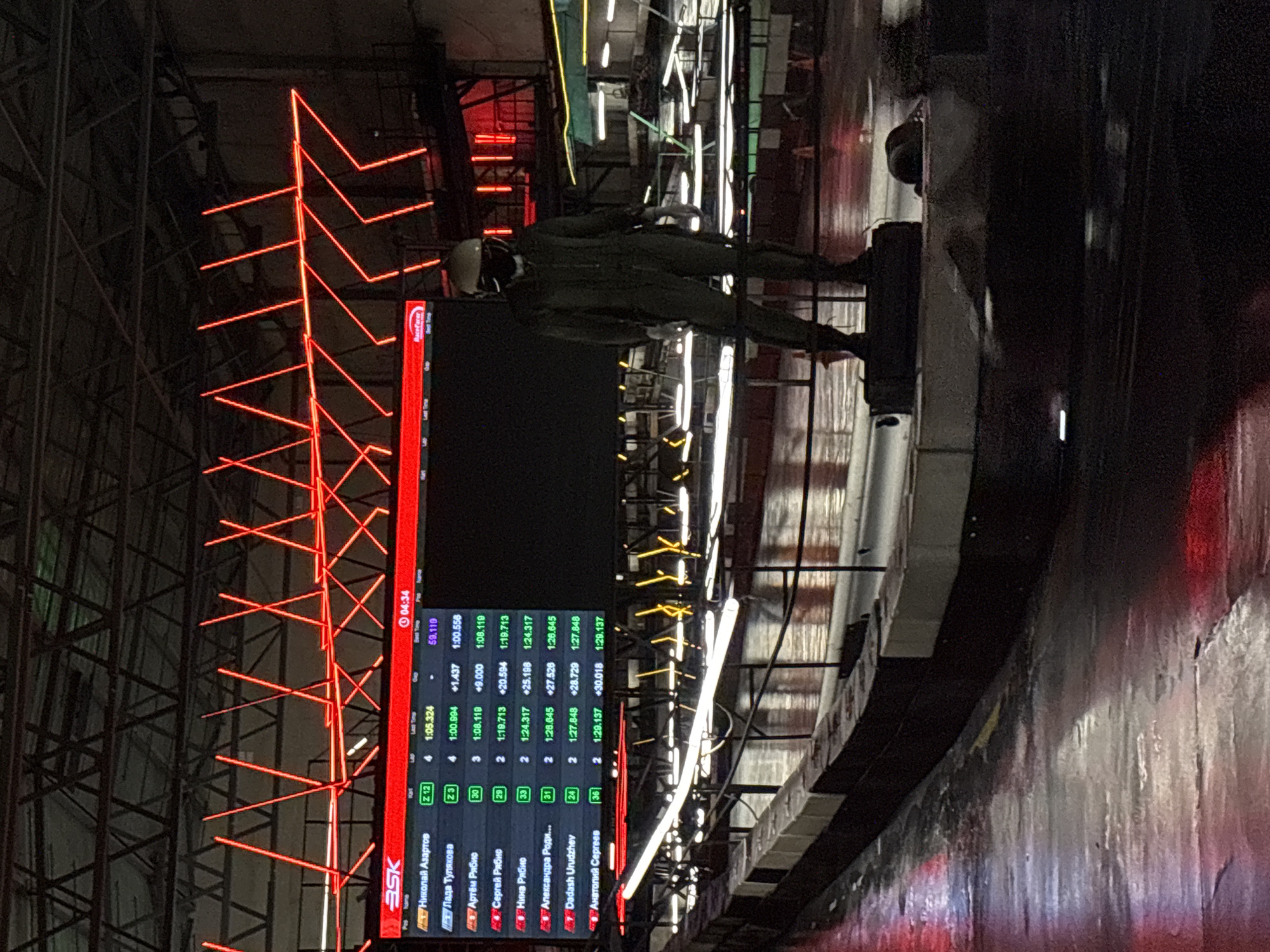 This media file captures a dimly lit, indoor scene, likely a sporting event or competition, with a distinct urban and industrial feel, possibly within Saint Petersburg, Russia. The primary subjects are a person in a dark uniform, possibly a participant or official, standing near a large digital scoreboard. The scoreboard displays race information, including participant names, lap times, and best times, suggesting a timed competition such as racing. The names on the scoreboard are in Russian, reinforcing the location context. The setting is characterized by exposed metal structures, neon red lighting accents forming geometric patterns, and bright white lights, creating a dramatic and atmospheric ambiance. The overall impression is one of a high-energy event, possibly taking place at night or in a space with controlled lighting. There are reflections on a wet or glossy surface in the foreground, suggesting the possibility of rain outside or a deliberately reflective floor. The person in uniform appears focused, standing alertly. The image's orientation is rotated, but the content remains discernible. Visible text includes "ASK" and "RaceFacer" on the scoreboard, along with participant names and race data.