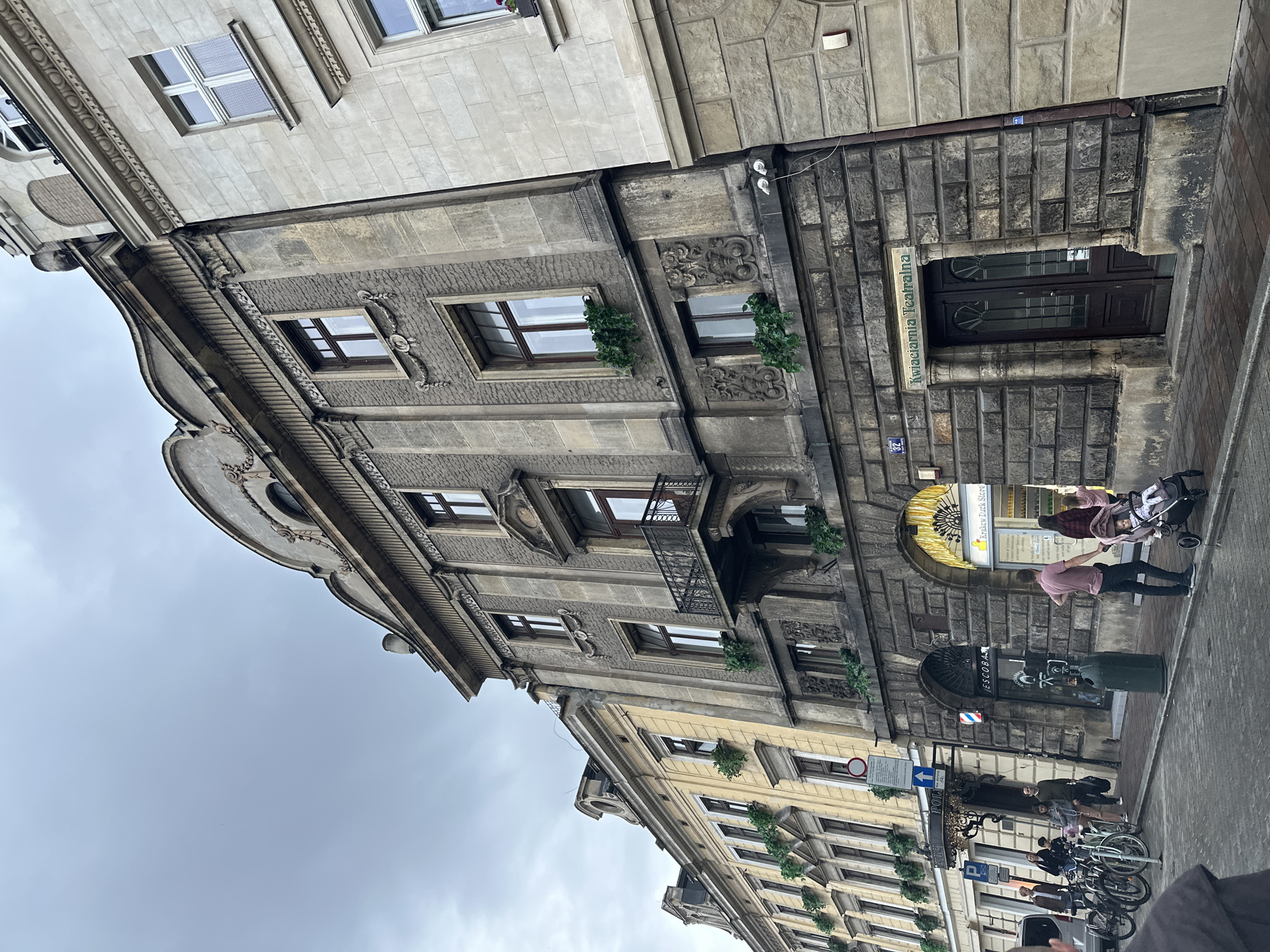 An exterior view of buildings on a street in Krakow, Poland, shows a cloudy sky overhead. The main building in the foreground features a weathered stone facade with decorative architectural elements, multiple windows, and a small balcony. A sign above a dark wooden door reads "Kwiaciarnia Teatralna," indicating a flower shop. To the left, another building with a lighter-colored facade and numerous windows is visible, with a row of bicycles parked along the sidewalk. People are walking on the sidewalk, including a person pushing a stroller with a child. The street is paved with cobblestones.