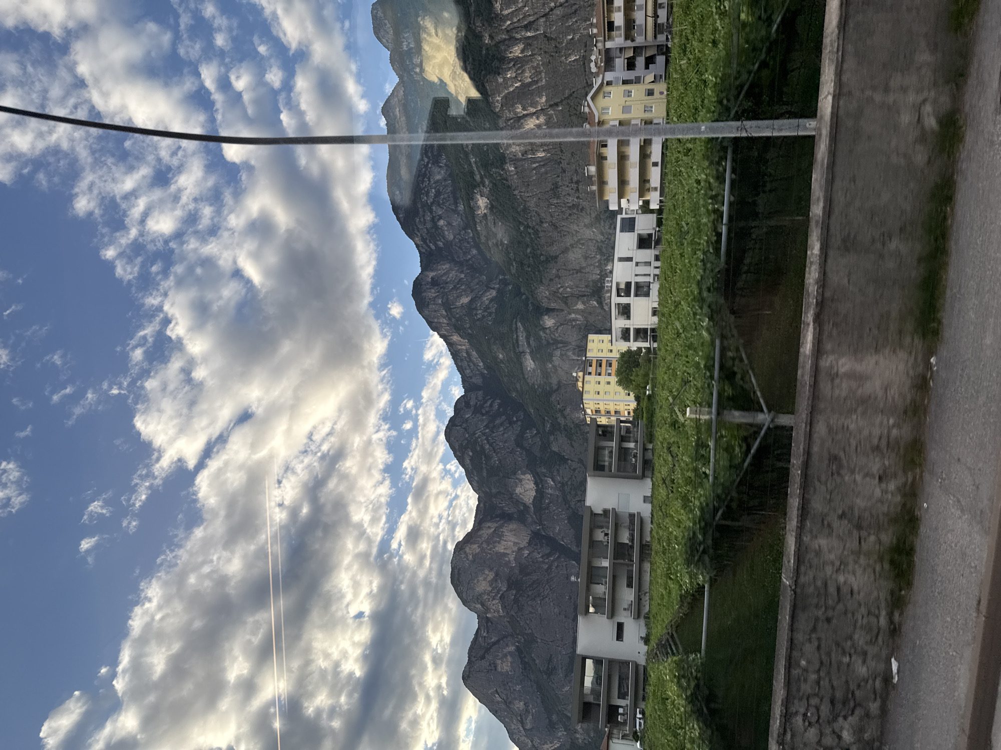 The image captures a scenic view of Mezzolombardo, Italy, dominated by a majestic mountain range in the background. The sky above is a canvas of blue adorned with dramatic clouds, with a faint contrail suggesting the passage of an aircraft. In the mid-ground, a modern apartment building with a white facade and multiple balconies stands out, bordered by lush green vineyards. Adjacent to it, a yellow apartment building adds a splash of color to the scene. A lamppost stands tall in the foreground, partially obscuring the view of a stone wall and a paved road below. The overall impression is one of a tranquil, picturesque town nestled against a breathtaking natural backdrop.