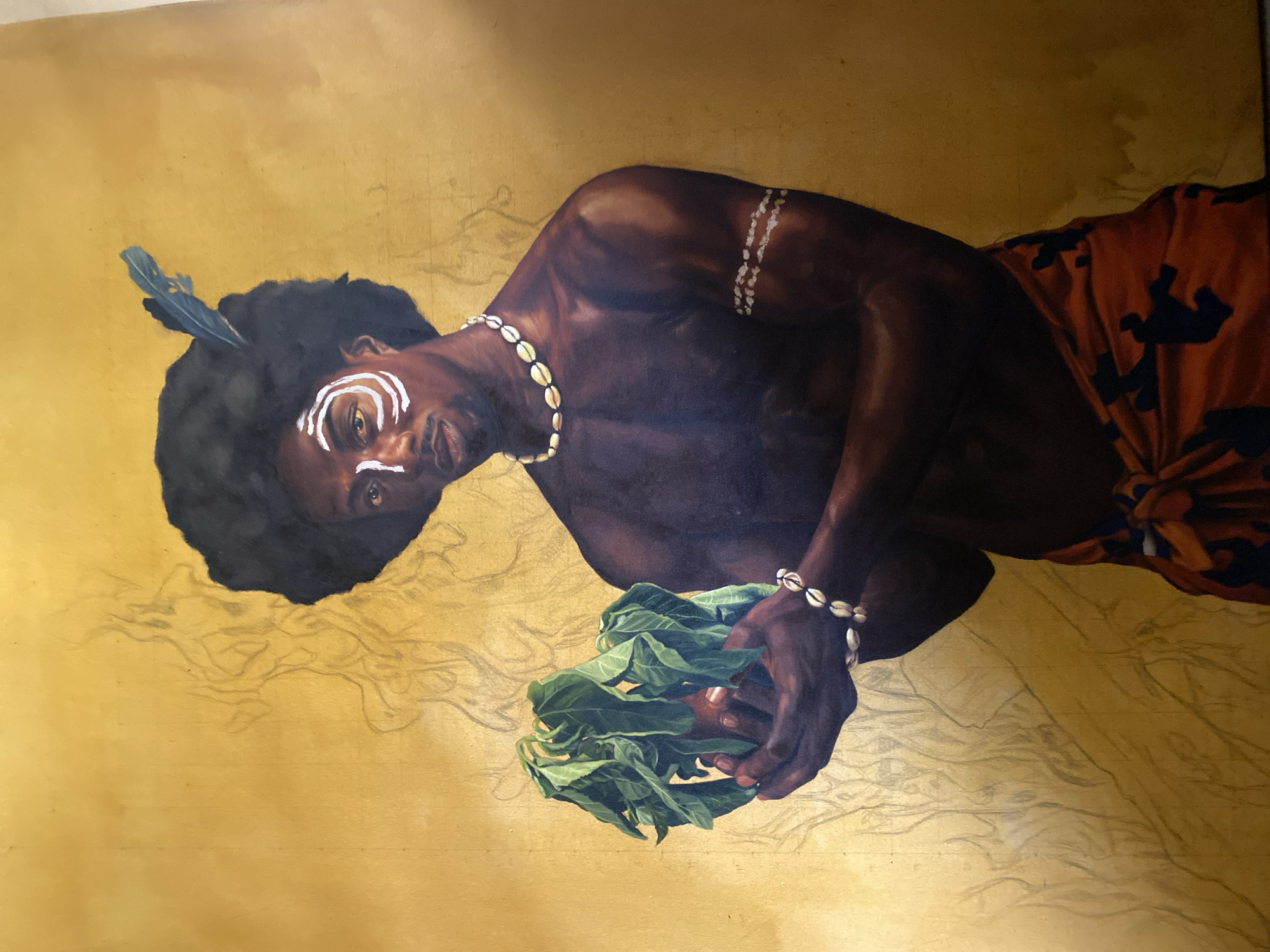 A painting depicts a shirtless, dark-skinned man with a feather in his hair and white markings on his face. He wears a necklace and bracelet made of shells and holds a cluster of green leaves in his hands. He is wearing a patterned cloth around his waist. The background is a gold color with faint pencil sketch marks. The location in the city of Ado, Nigeria is not evident from this image.