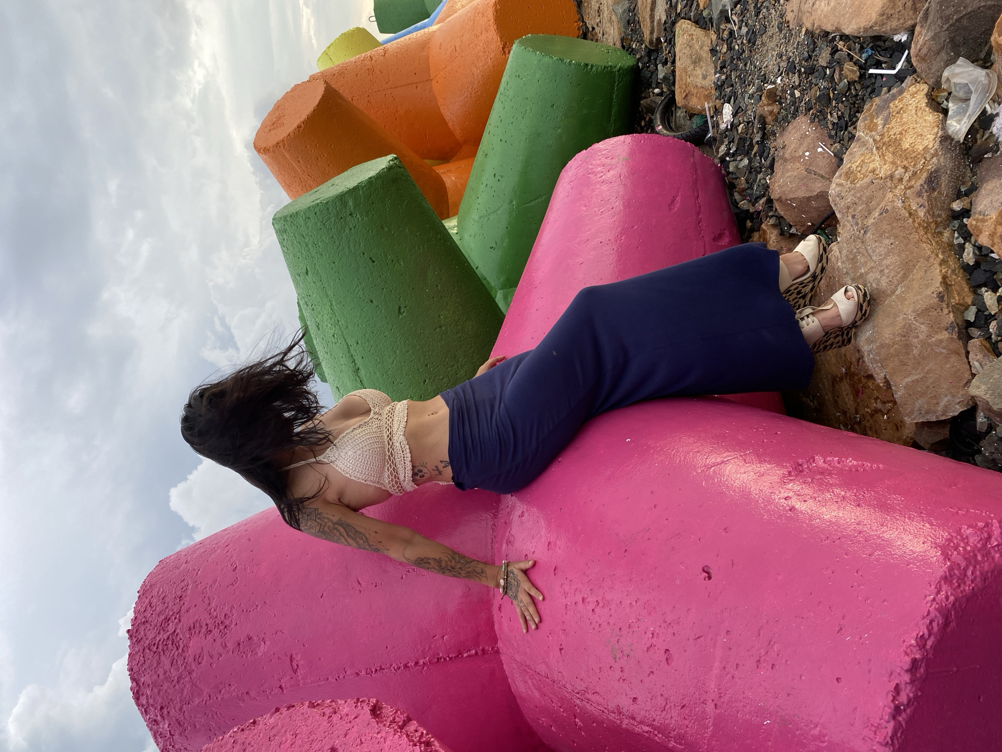 An outdoor, full-length shot shows a woman posing on large, brightly colored concrete blocks, likely breakwater or coastal defense structures, under an overcast sky. The woman is positioned horizontally across a vibrant pink block. She has long, dark hair that appears windswept and wears a light-colored, crocheted crop top that reveals her midriff, along with dark blue wide-leg pants or a skirt. Distinctive tattoos are visible on her left arm and shoulder, extending towards her back, as well as smaller designs on her torso. She is wearing light-colored wedge sandals with a leopard print design.
Her left arm is extended along the pink block, with her hand resting on its surface. In the background, other concrete blocks are visible, painted in green, orange, and hints of yellow. The ground to the right of the woman is rocky and uneven, featuring a mix of larger brown rocks and smaller pebbles, along with some scattered debris including a plastic bag and white plastic pieces. The sky above is largely covered with light grey clouds. The scene suggests a coastal or waterfront setting in Phu Thuy Ward, Vietnam. There is no visible text in the image.