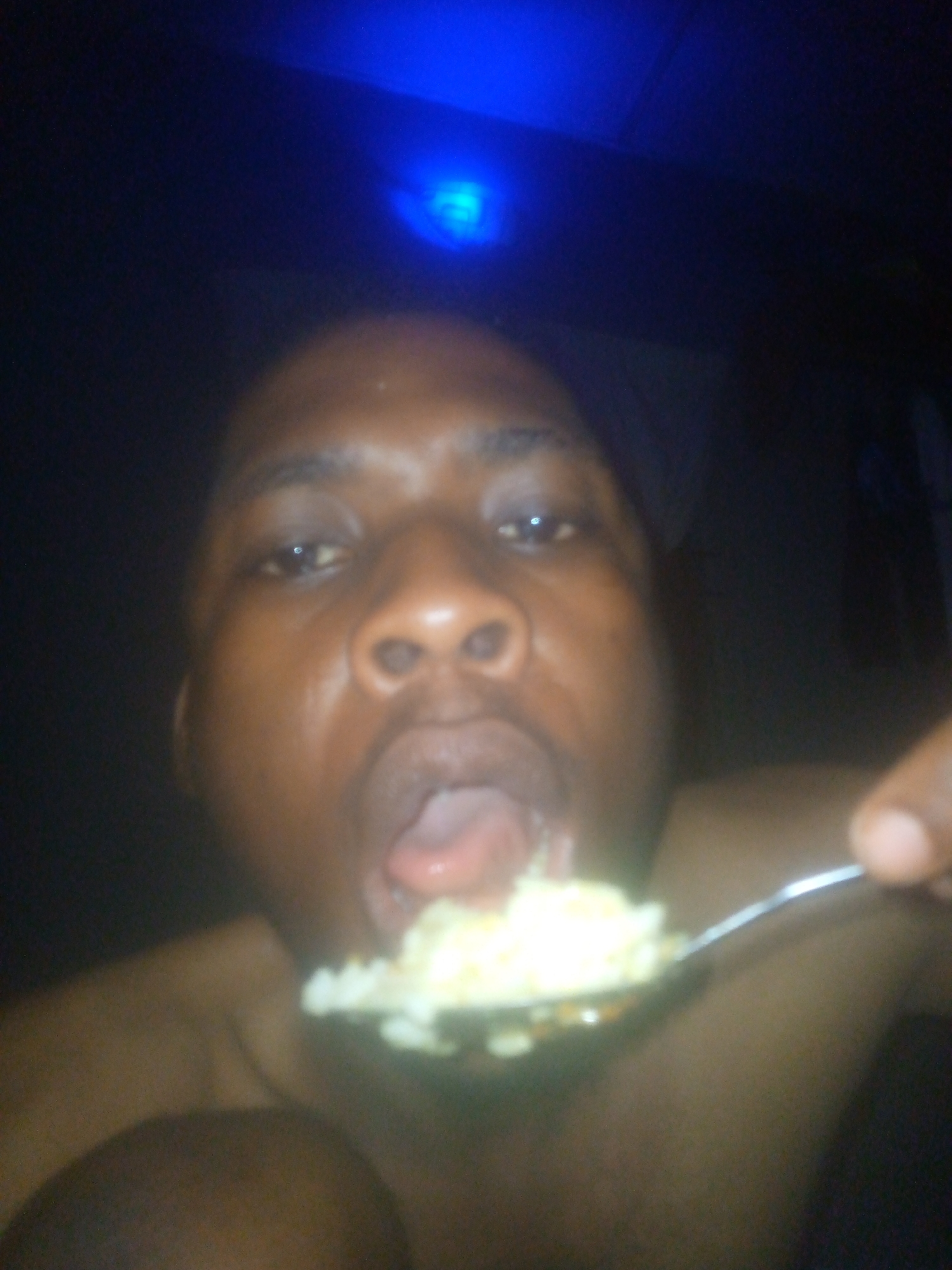 A close-up shot shows a person with dark skin about to eat a spoonful of yellow food. The person's mouth is open, and their tongue is visible. Above them is a blue light. The background is dark and out of focus. The location is Unknown, Unknown.