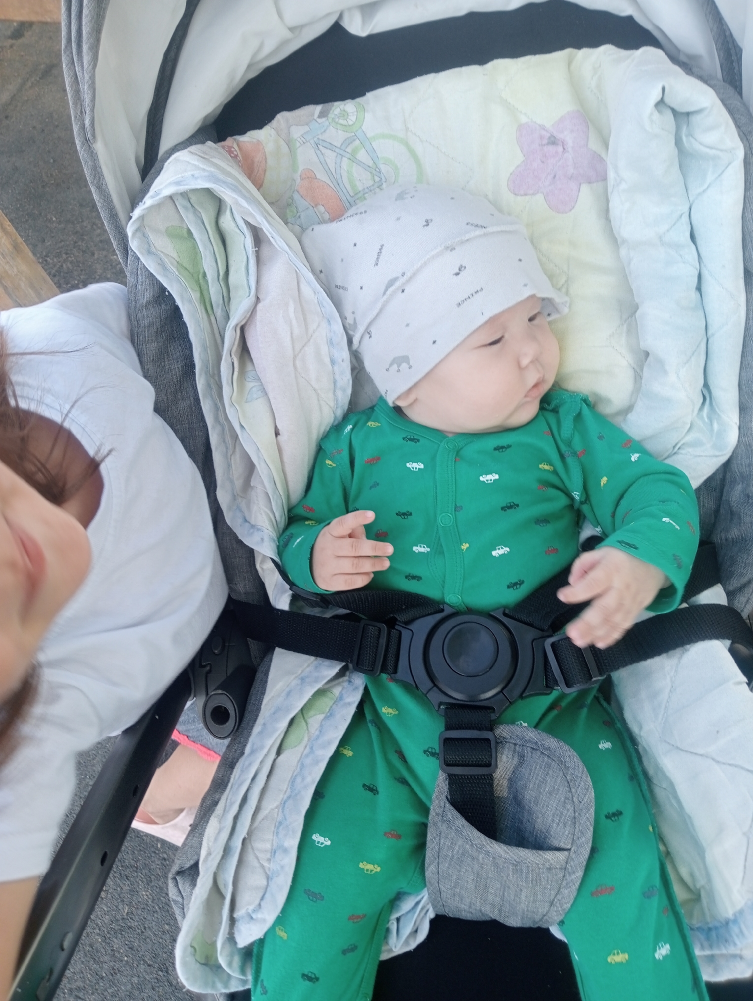 A baby wearing a green onesie with a car pattern and a white hat with elephant patterns is lying in a stroller. The stroller is lined with light-colored blankets, one of which has a pink flower pattern. A person's arm and head, covered with dark hair, are visible on the left side of the image, suggesting they are either holding or attending to the baby. The background appears to be an outdoor setting with a paved or gravel surface and some greenery, possibly a park or street in Aksu, Kazakhstan. The stroller has a black safety harness with a large buckle.