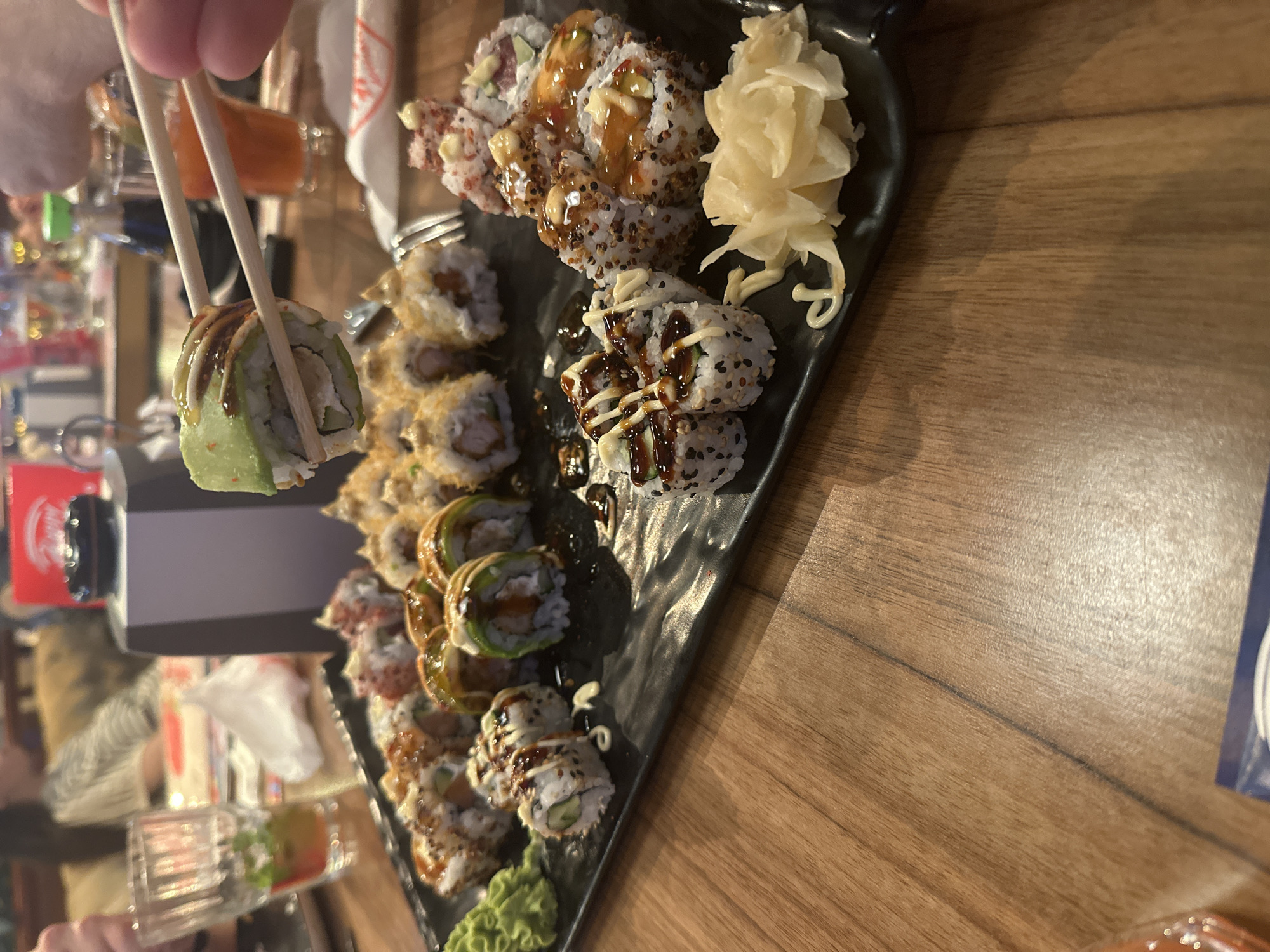 The image captures an indoor dining scene within a restaurant in Unknown, Unknown. The primary subject is a large, black, rectangular serving platter, textured and matte, holding an extensive assortment of sushi rolls. This platter rests on a light-brown wooden table with a visible grain.
A human hand, visible in the upper right quadrant of the image, utilizes light-colored wooden chopsticks to elevate a single sushi roll from the platter. This specific roll is topped with thinly sliced avocado and liberally drizzled with a dark, viscous sauce, likely a tare or eel sauce. Its cross-section reveals rice, a white creamy filling, and a green interior component, likely cucumber.
The platter itself displays a variety of other sushi preparations:
- Numerous rolls coated with black and white sesame seeds, some also adorned with drizzles of white, creamy sauce and darker sauces.
- Rolls with a crispy, golden-brown topping.
- On the far left of the platter, a distinct portion of green wasabi paste is visible.
- On the far right, a mound of pale yellow pickled ginger is presented, alongside a decorative swirl of white, mayonnaise-like sauce.
In the blurred background, several individuals are seated, suggesting an active dining establishment. A clear drinking glass containing a beverage with a green garnish is partially visible on the table to the left. To the right, behind the chopsticks, a dark, small bottle (consistent with a soy sauce dispenser) and a glass holding an orange-colored drink are discernible. On a distant table in the background, a dark, multi-faceted object with a black top, possibly a call button or device, is also present. The overall lighting is warm and artificial, indicative of an indoor setting.