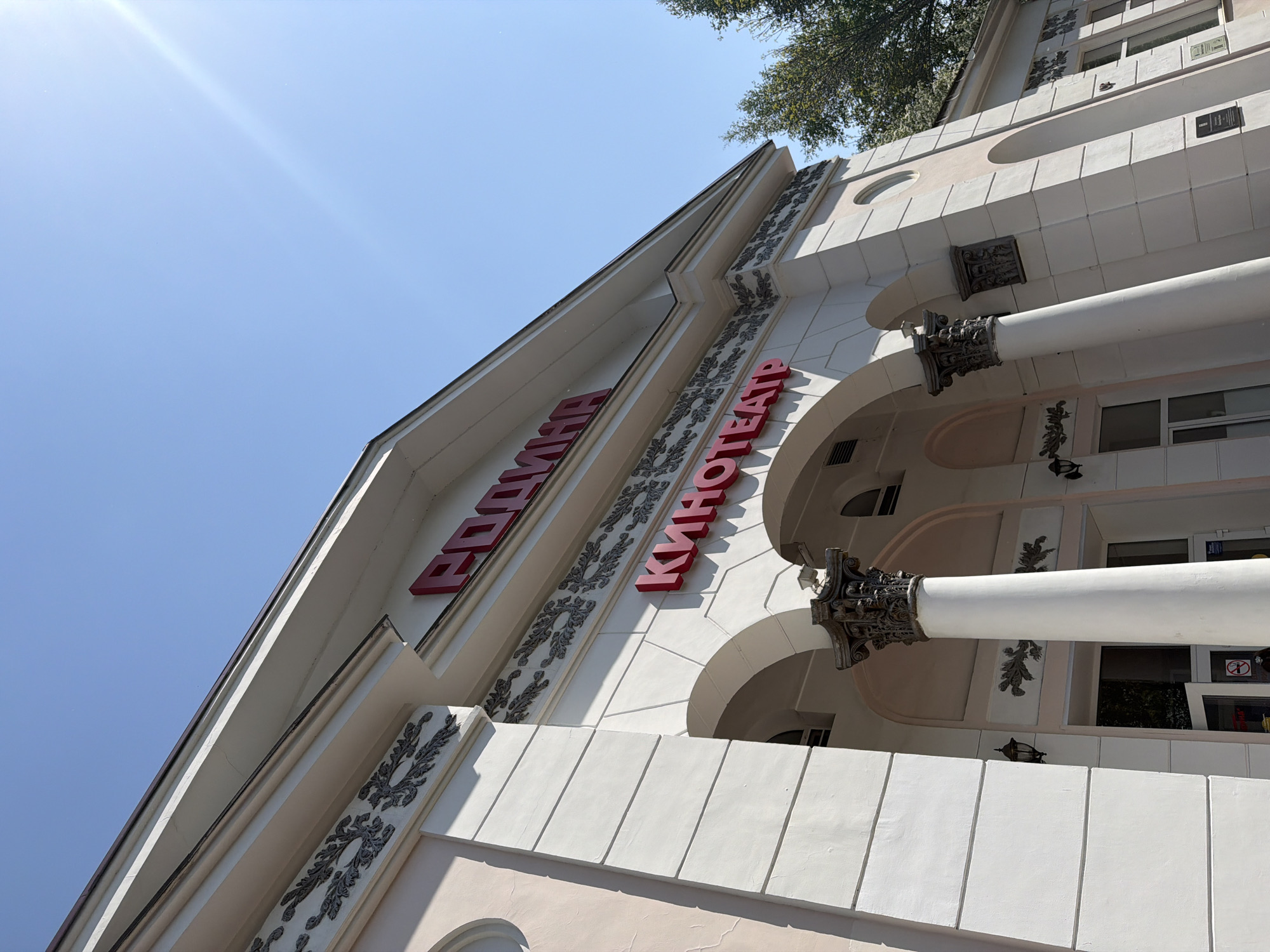 The image shows the facade of a building in Birobidzhan, Russia, identified by the Cyrillic lettering "ЕДИНАЯ КИНОТЕАТР," which translates to "United Cinema." The building features a classical architectural style, with decorative moldings, columns, and arches. The exterior is painted in light colors, including white and beige. The sign is in red. The sky is visible in the background, and part of a tree is visible.