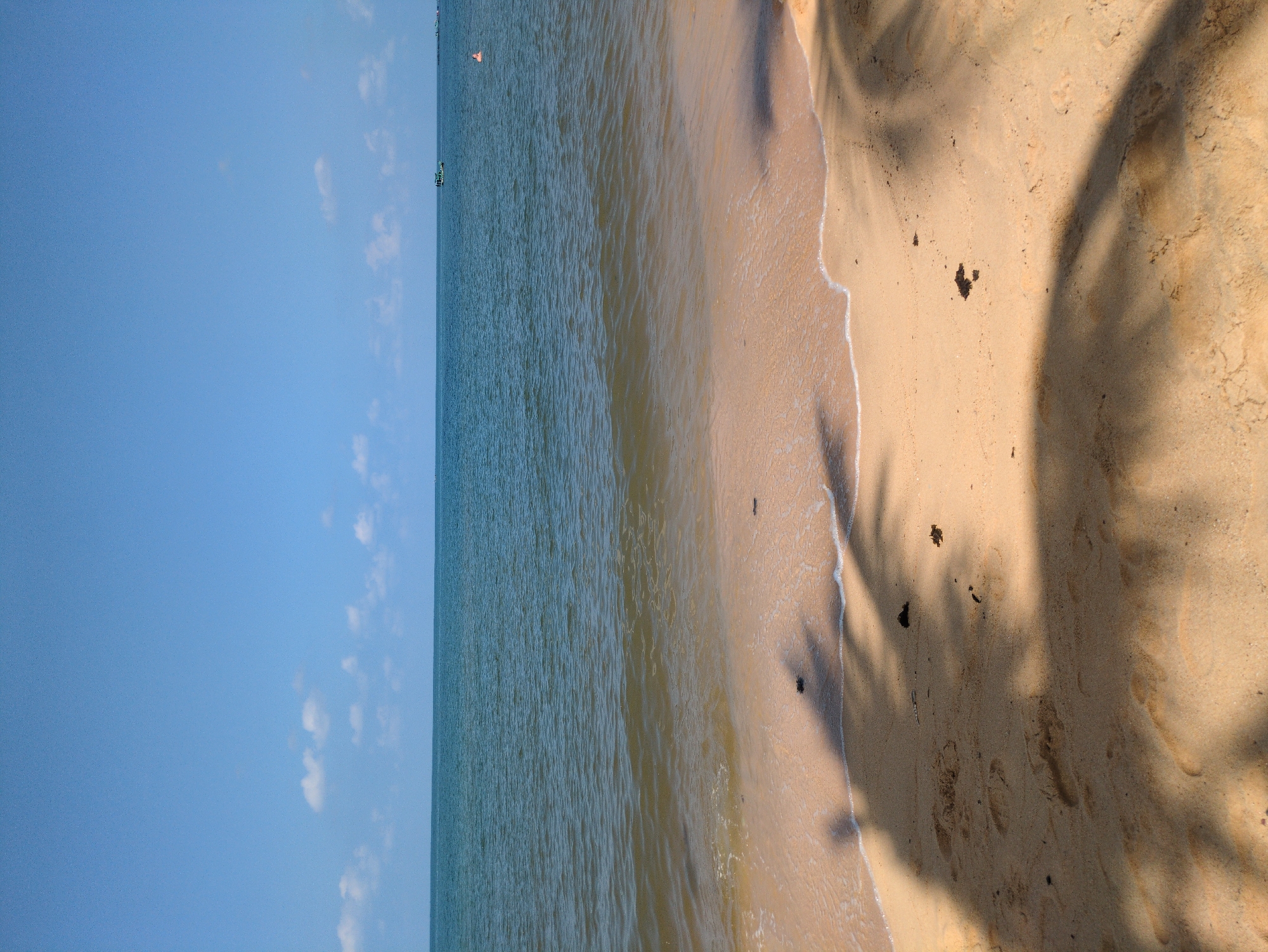The image shows a beach scene in Phú Quốc, Vietnam. The sky is blue with scattered clouds, transitioning to the ocean, which is calm with gentle waves lapping the shore. The sandy beach is tan and exhibits shadows from palm trees. The image does not show any people or specific objects, but the presence of a boat can be seen in the distance.