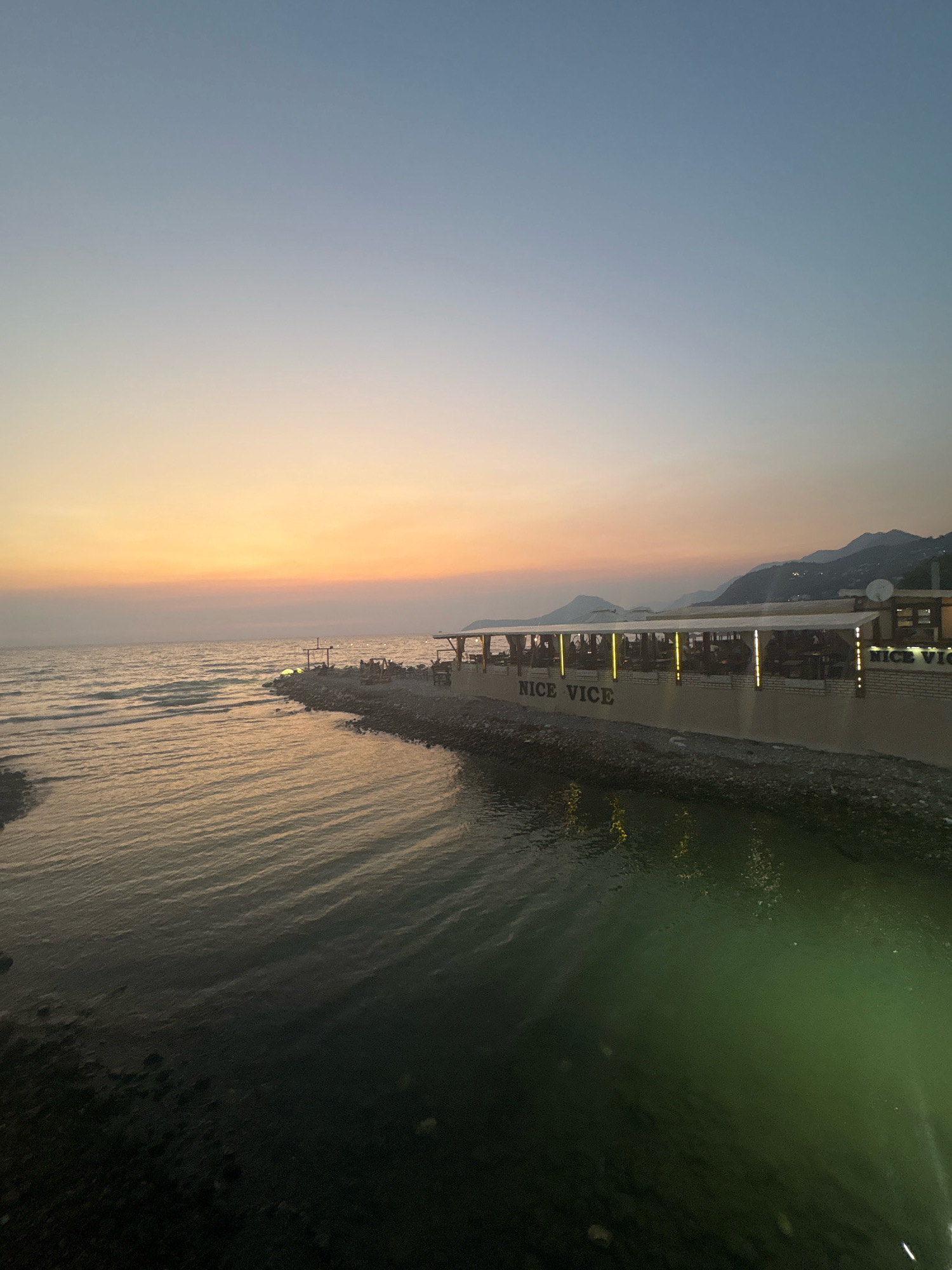 The image shows a waterfront scene at dusk in Bar, Montenegro. A long, low building with a covered outdoor seating area runs along the shore. The building has "NICE VICE" written on its facade in large letters, illuminated by overhead lights. Tables and chairs are visible under the cover, suggesting it is a restaurant or bar. Beyond the building, mountains rise in the background. The sea is calm with gentle waves, reflecting the soft colors of the sunset in the sky, which transitions from pale yellow and orange at the horizon to a light blue at the top. The shore appears to be made of pebbles. No people are clearly identifiable.