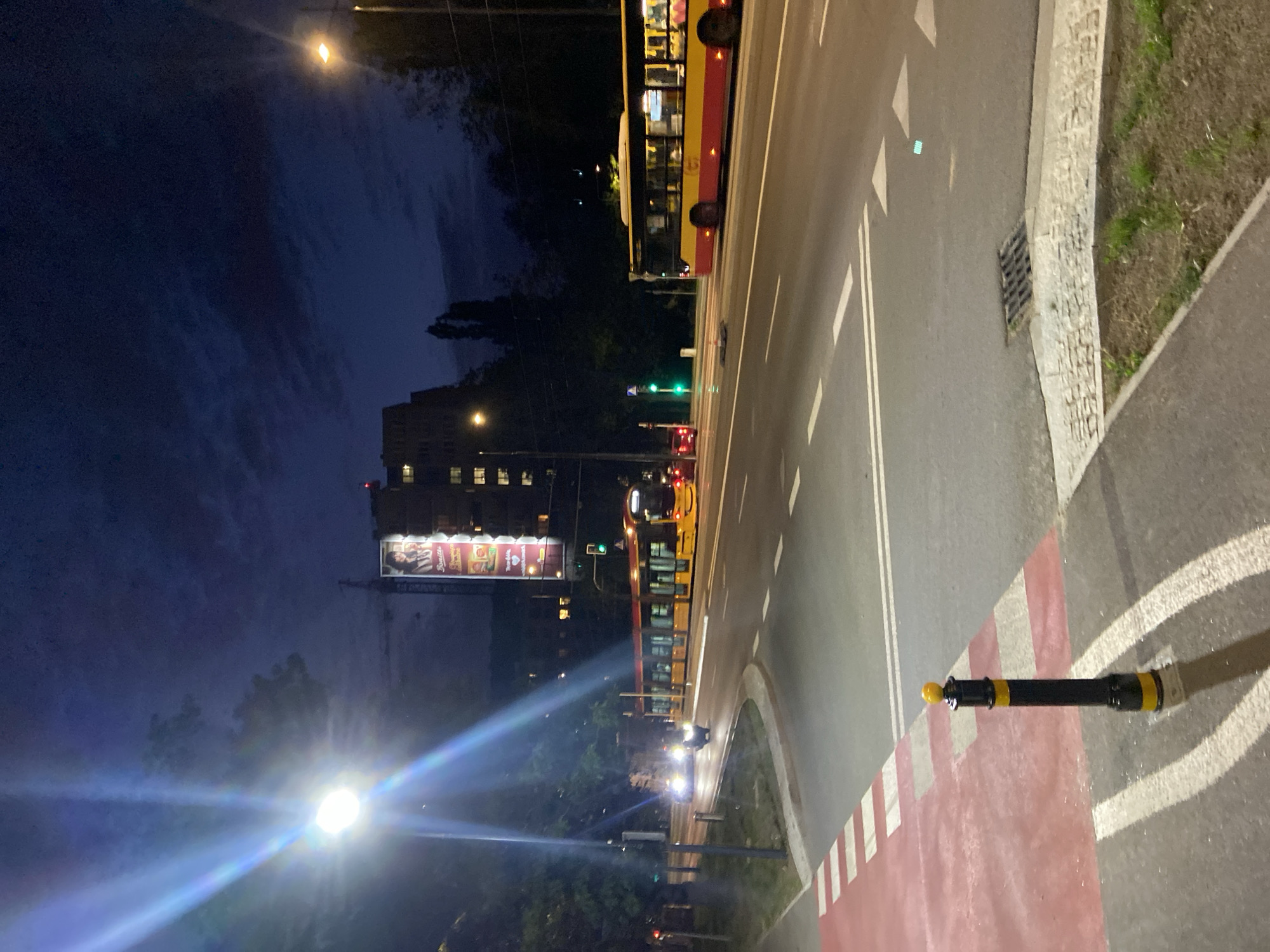 The image captures a nighttime street scene in Warsaw, Poland, likely at an intersection. A yellow and red tram is visible moving to the left, while a similar colored bus travels to the right on a multi-lane road. Streetlights illuminate the scene, with one particularly bright one in the foreground casting a strong glare. Buildings and trees line the background, and a large billboard with an advertisement is mounted on a building. The road features crosswalks, lane markings, and a traffic light showing green. A black and yellow bollard stands on a red-painted pedestrian crossing. The sky is dark with some clouds.