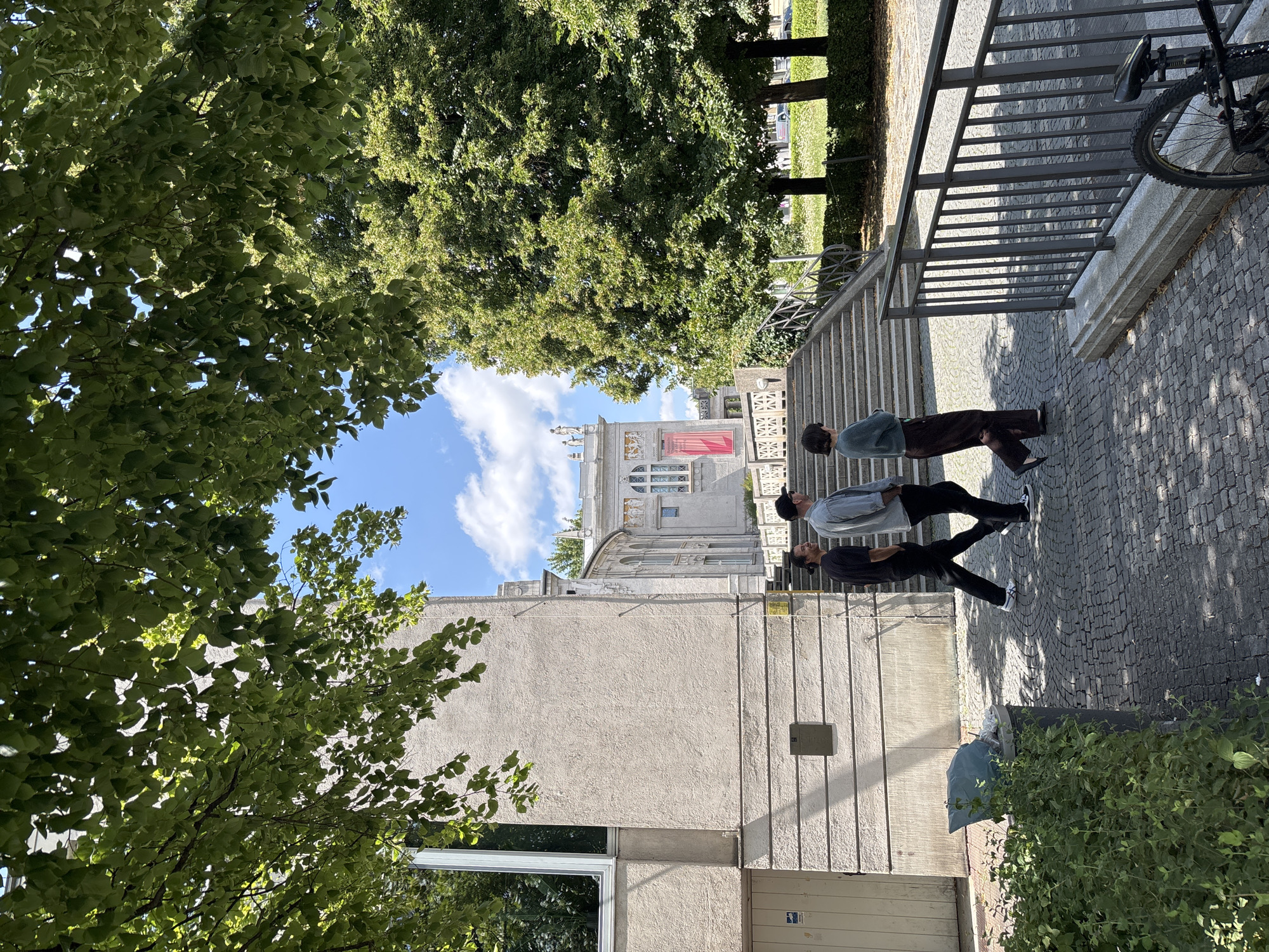 Three people are walking on a cobblestone path next to a building and a set of stairs. The building is light grey with horizontal banding on the lower section and a large window on the left. A red banner is hanging on the facade of a more ornate building in the background. Large, leafy green trees are prominent in the foreground and to the right. The sky is blue with scattered white clouds. A bicycle is parked to the right of the image, and a trash receptacle with a light blue bag is to the left, partially obscured by greenery. The scene appears to be in an urban park or public space. This image was taken in front of the "Altes Rathaus" (Old Town Hall) in Munich, Germany.