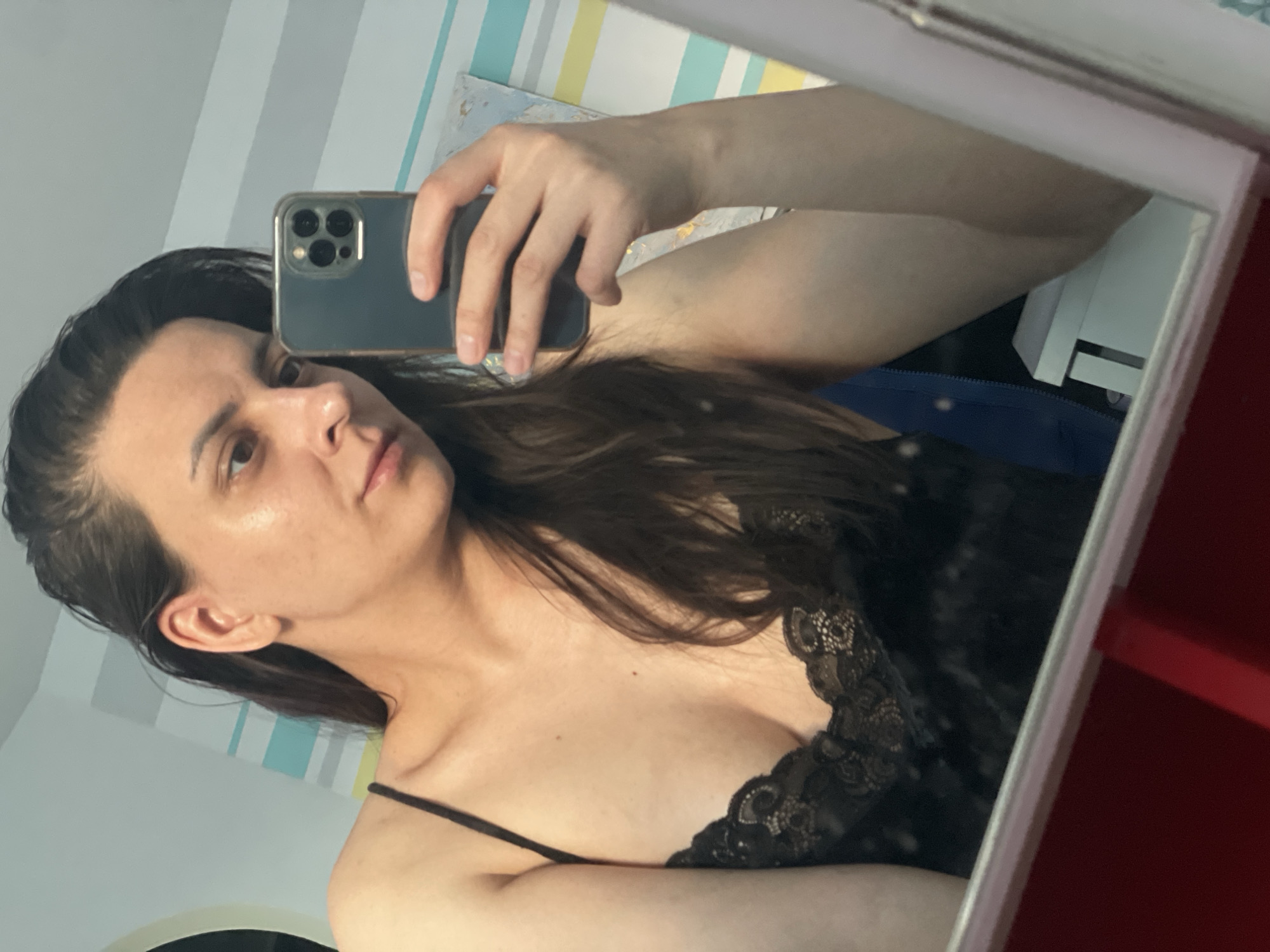 A woman is taking a selfie in a mirror. She is holding a smartphone, likely an iPhone, with a dark-colored case. The woman has dark hair and is wearing a black lace top with thin straps. The background includes a wall with pastel-colored stripes, a blurred painting, and a glimpse of furniture, possibly a desk or cabinet. The mirror has a frame, parts of which appear to be red. The location is Unknown, Unknown.